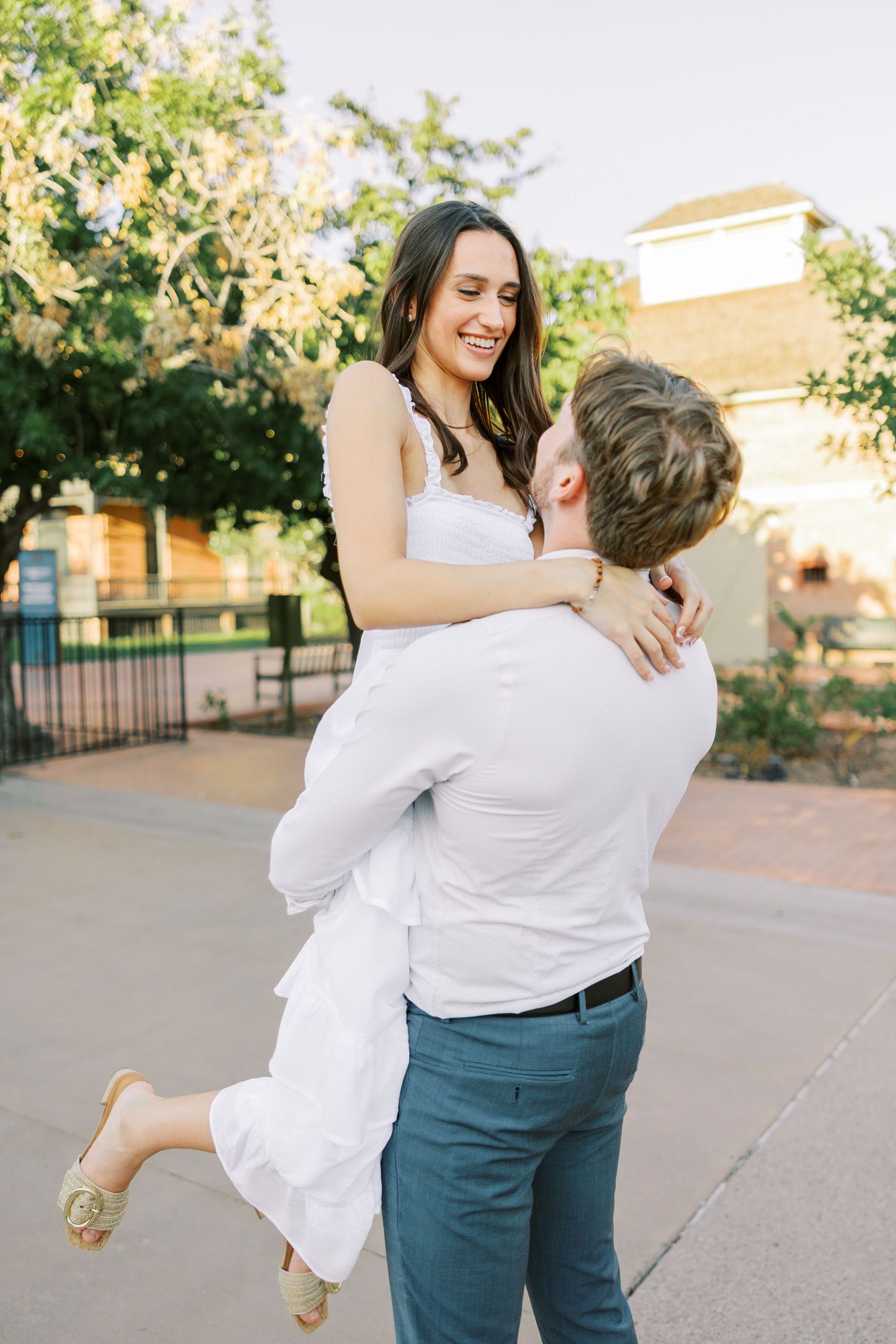 Couple in white and blue outfits dancing playfully outdoors with trees in background.