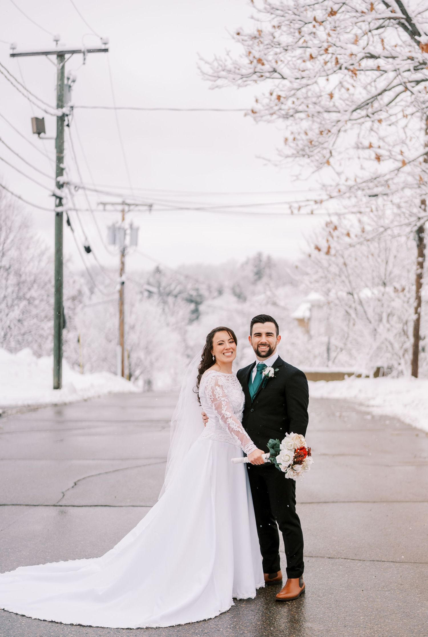 Newlyweds pose playfully on a winter street with snow-covered trees in the background.