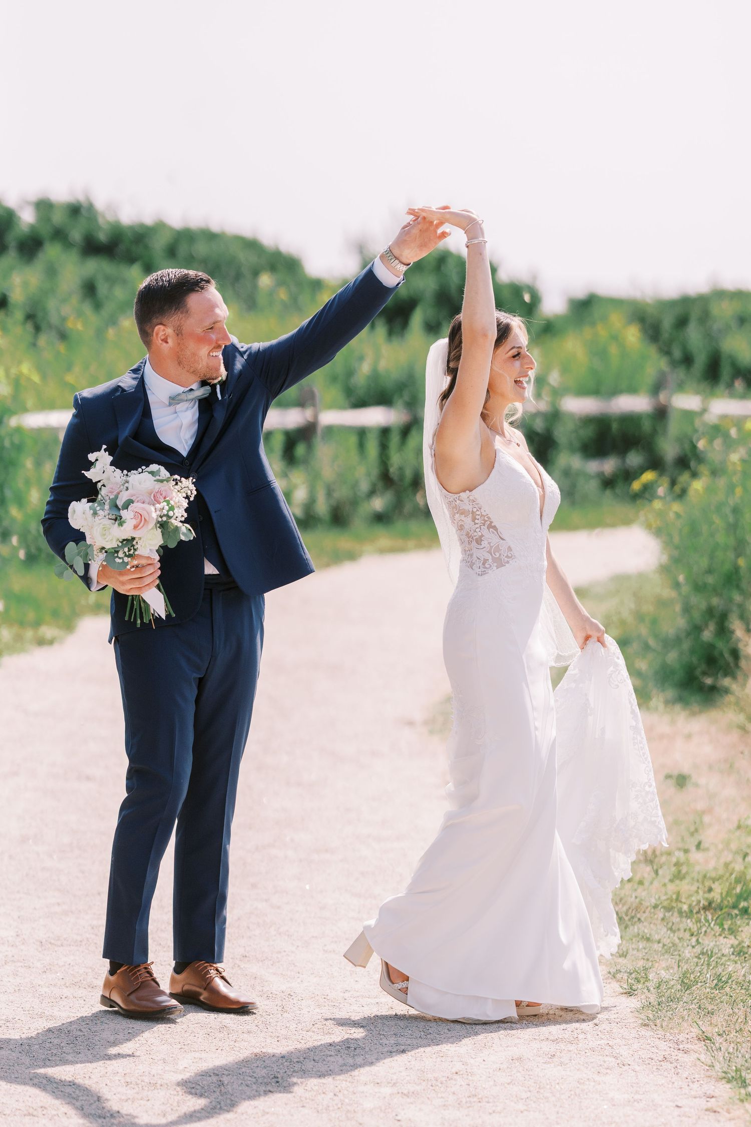 A couple in wedding attire dance together on a path outdoors, with the bride holding a pink and white bouquet.