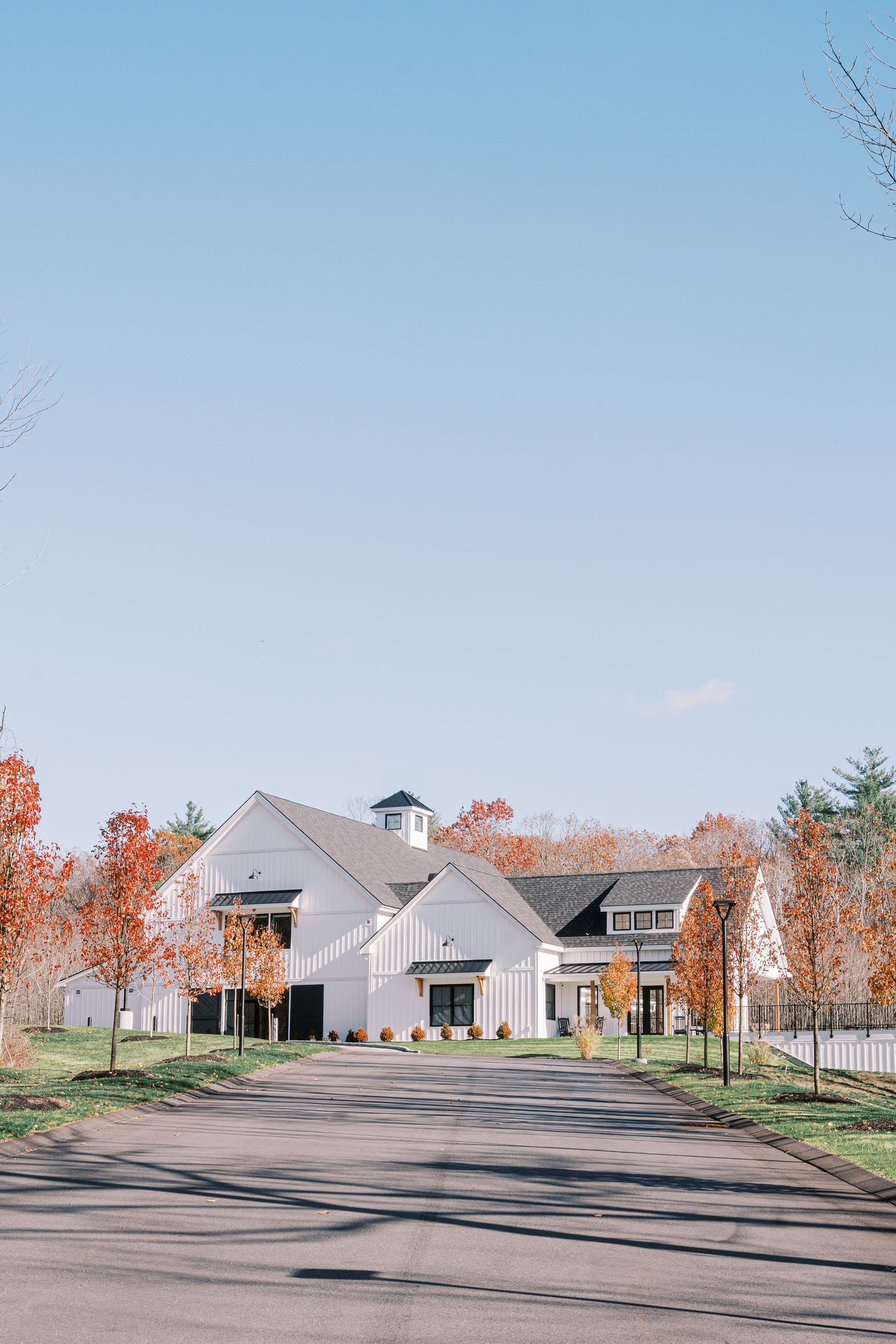 Modern white farmhouse-style buildings with black trim set against autumn trees and a clear blue sky in a rural setting.