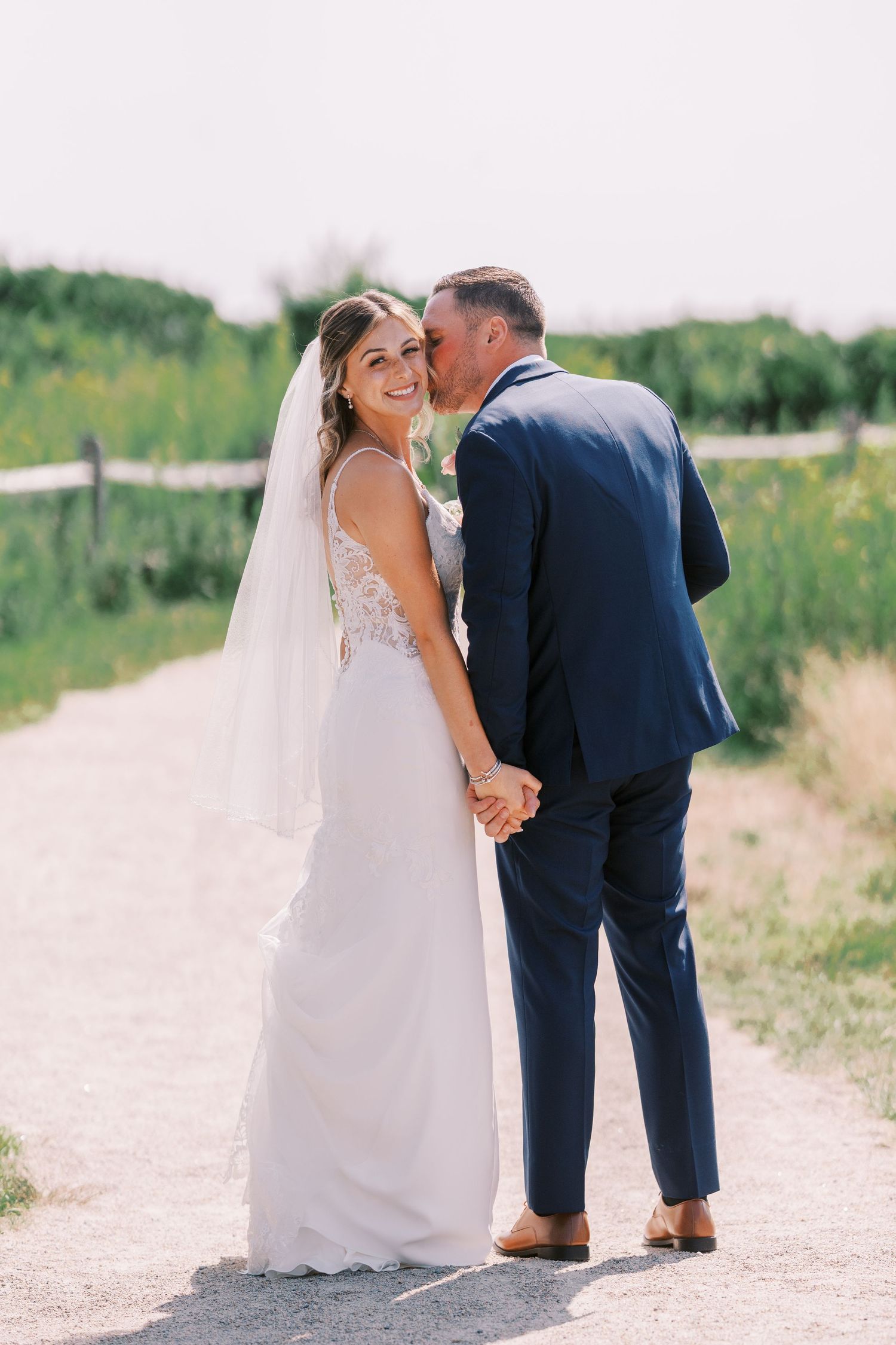 A couple in wedding attire walks along a dirt path surrounded by green fields on a sunny summer day.