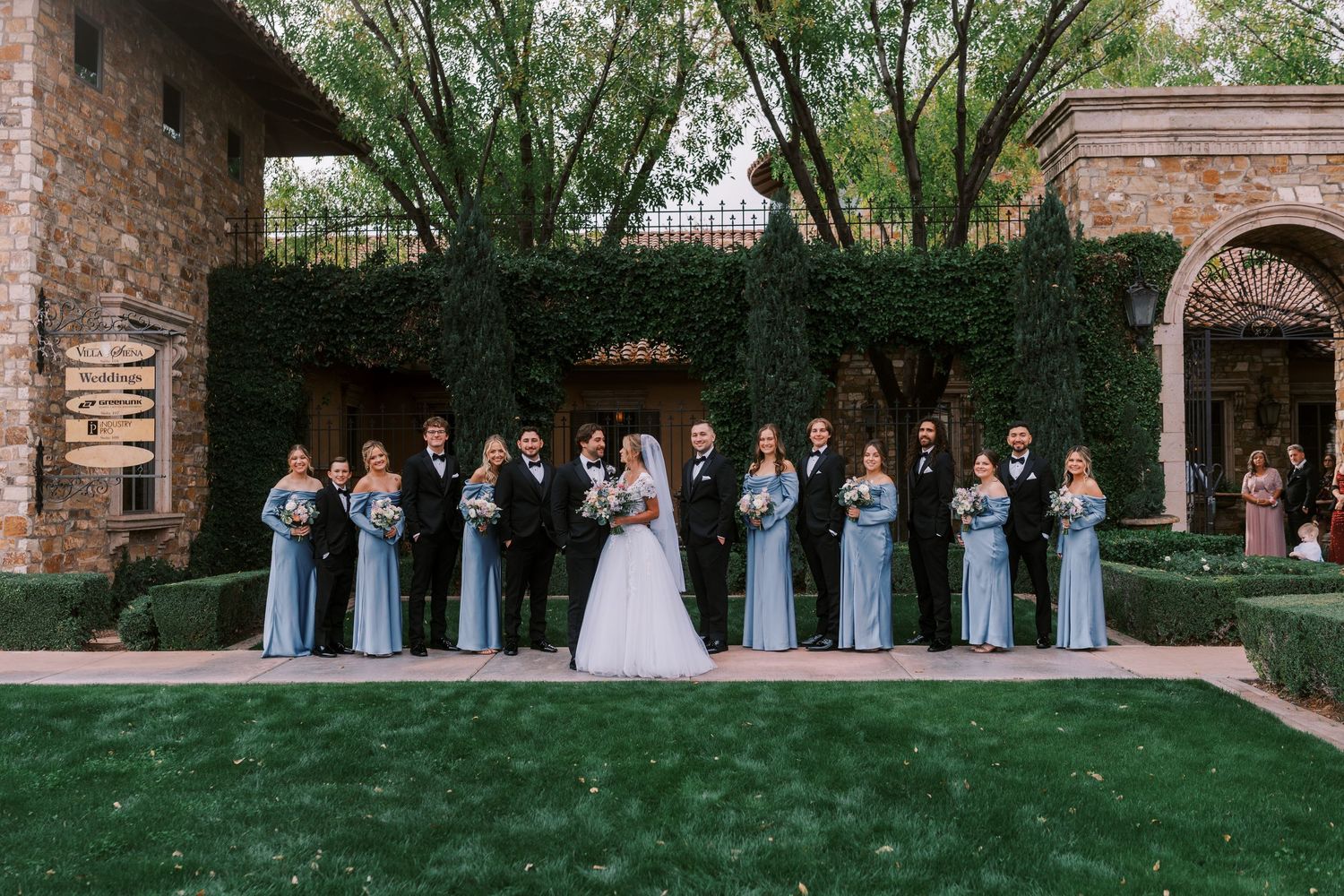Wedding party stands in elegant formation along a rustic brick archway during an outdoor ceremony.