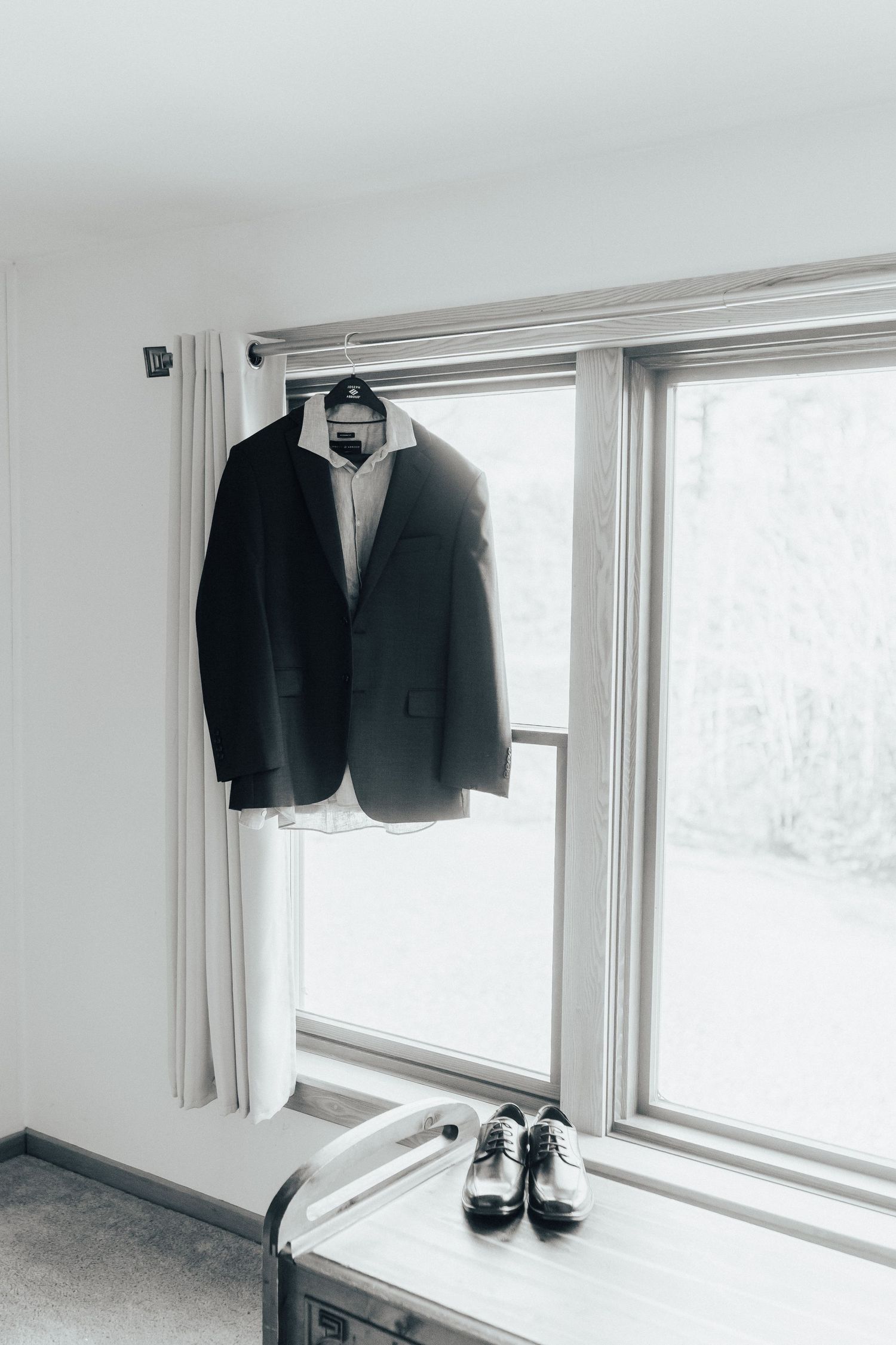 Black suit jackets and dress shoes displayed in minimalist white room settings against windows with natural light.