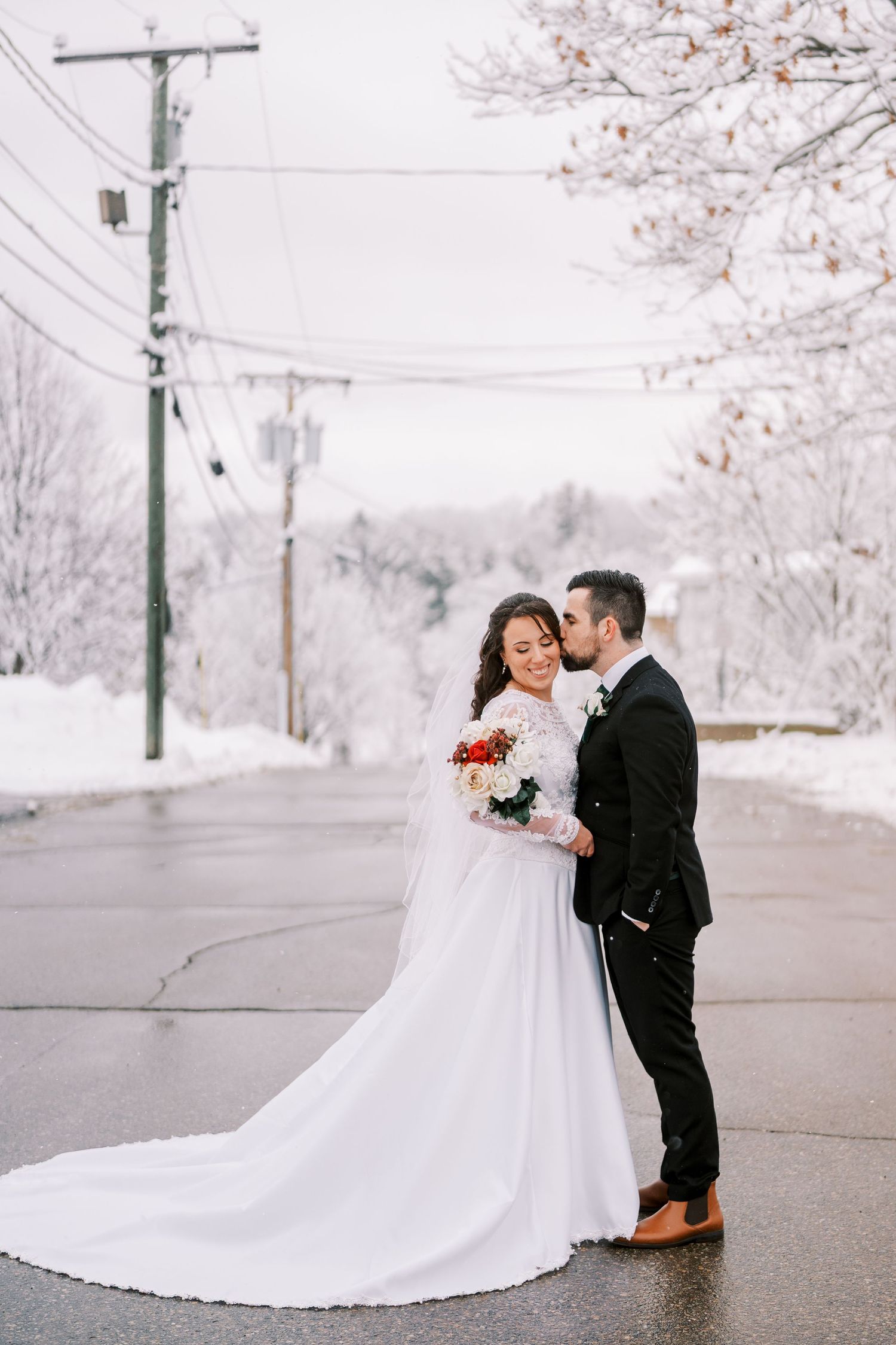 Newlyweds share an intimate moment on a snowy street under cherry blossom trees.