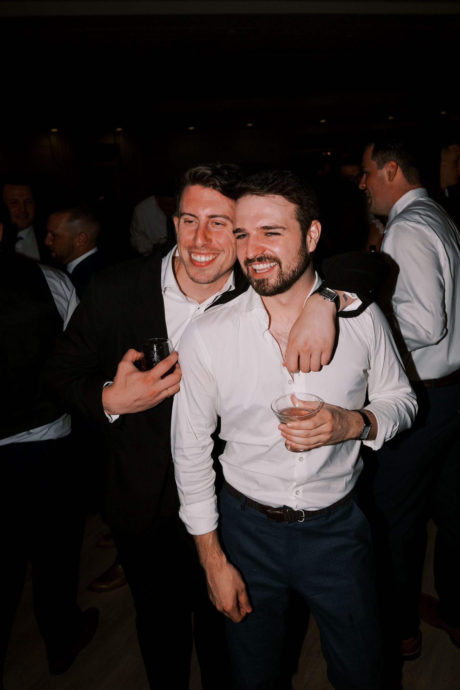 Two people in white shirts and dark pants embrace while dancing and laughing at a dimly lit event.