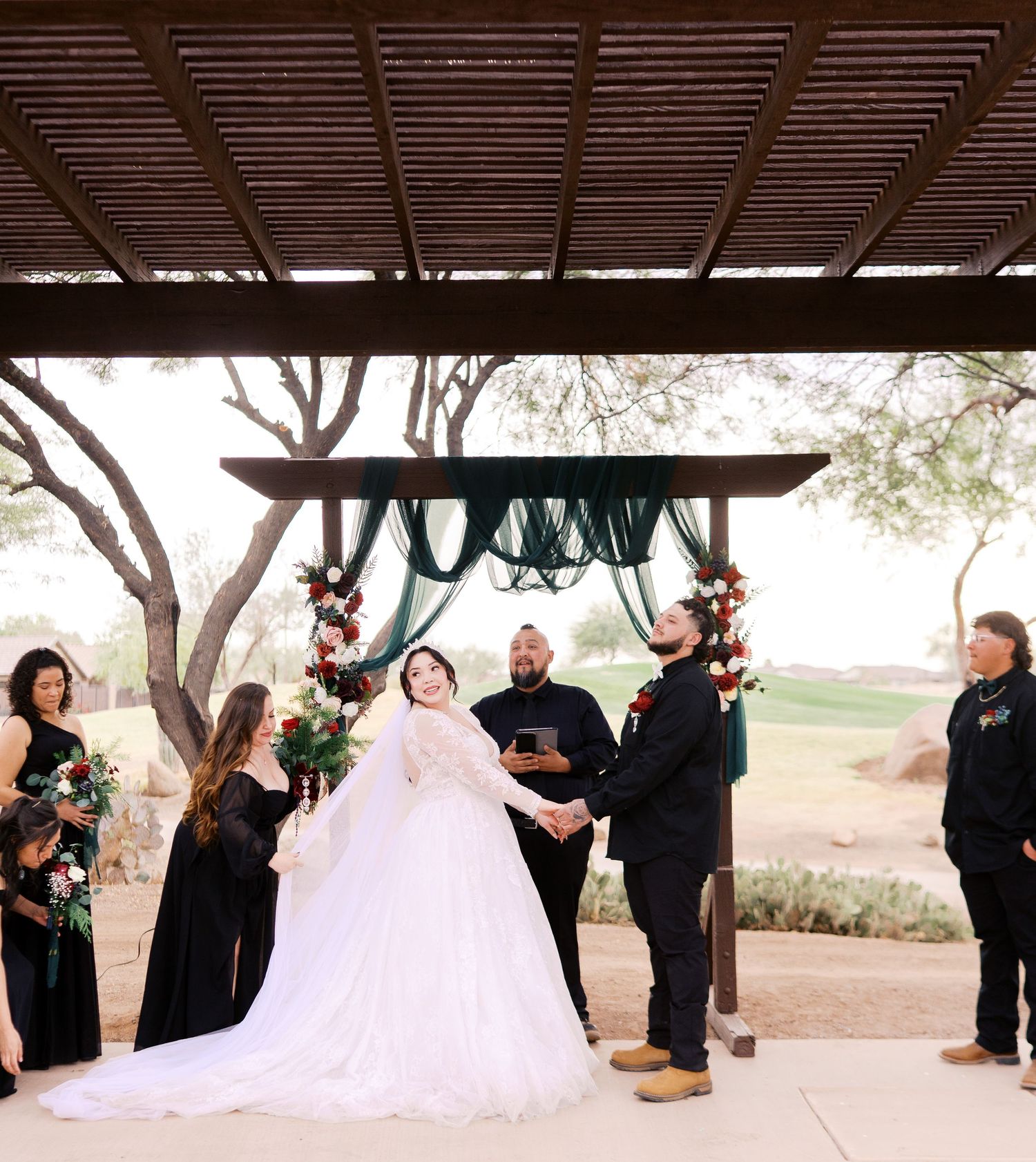An outdoor wedding ceremony takes place under a wooden pergola with attendants in black and a flowing white gown.