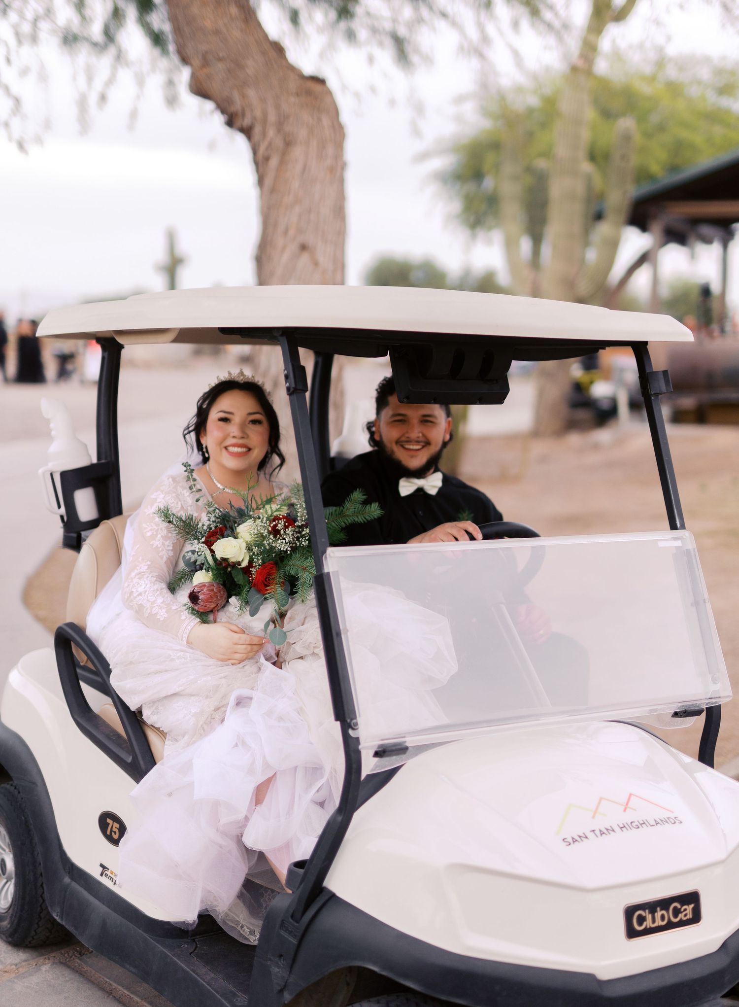 Wedding couple smiles while sitting in a white golf cart together at an outdoor venue with trees in background.