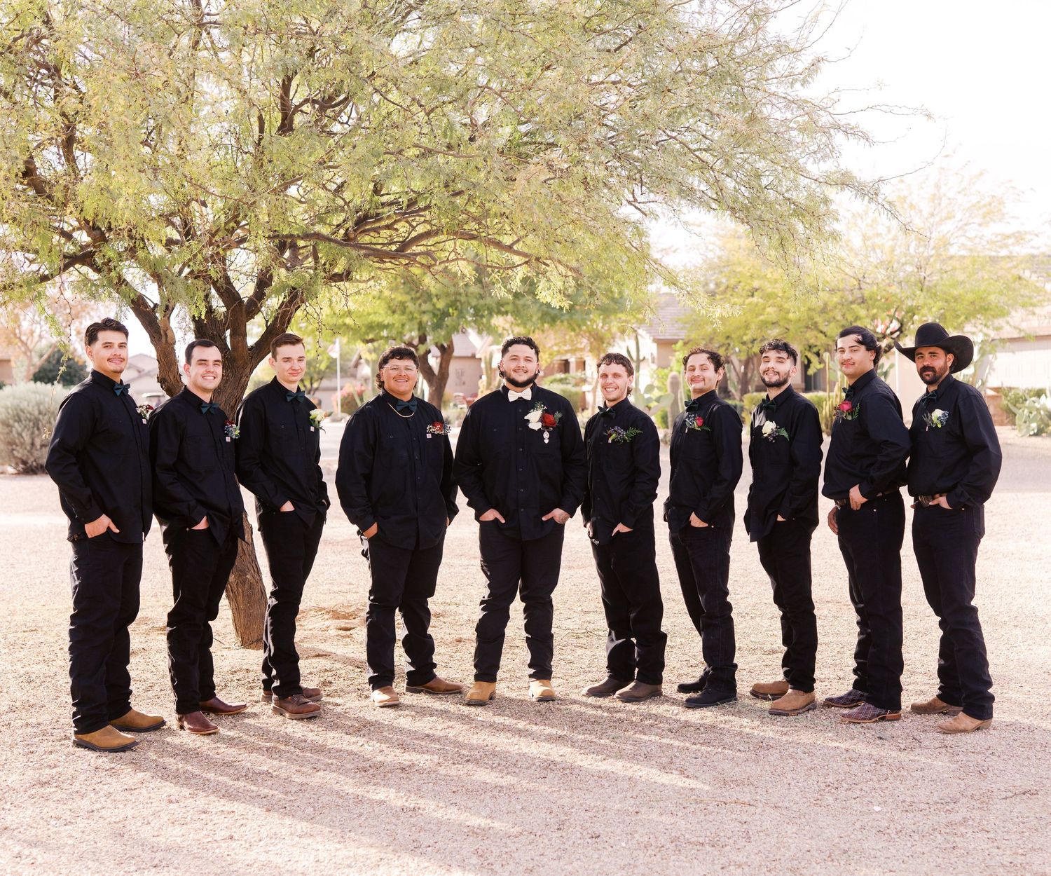 A wedding party in matching black attire with brown boots stands in a line outdoors beneath a large desert tree.