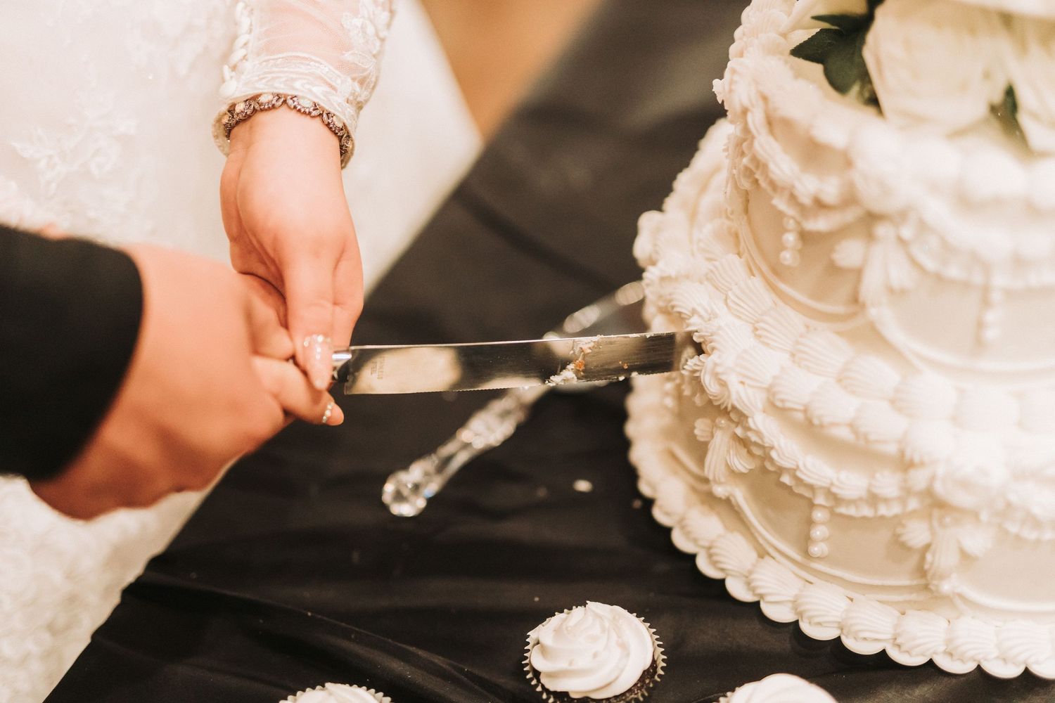Close-up view of hands holding a knife cutting into a white tiered wedding cake during the traditional cake cutting ceremony.