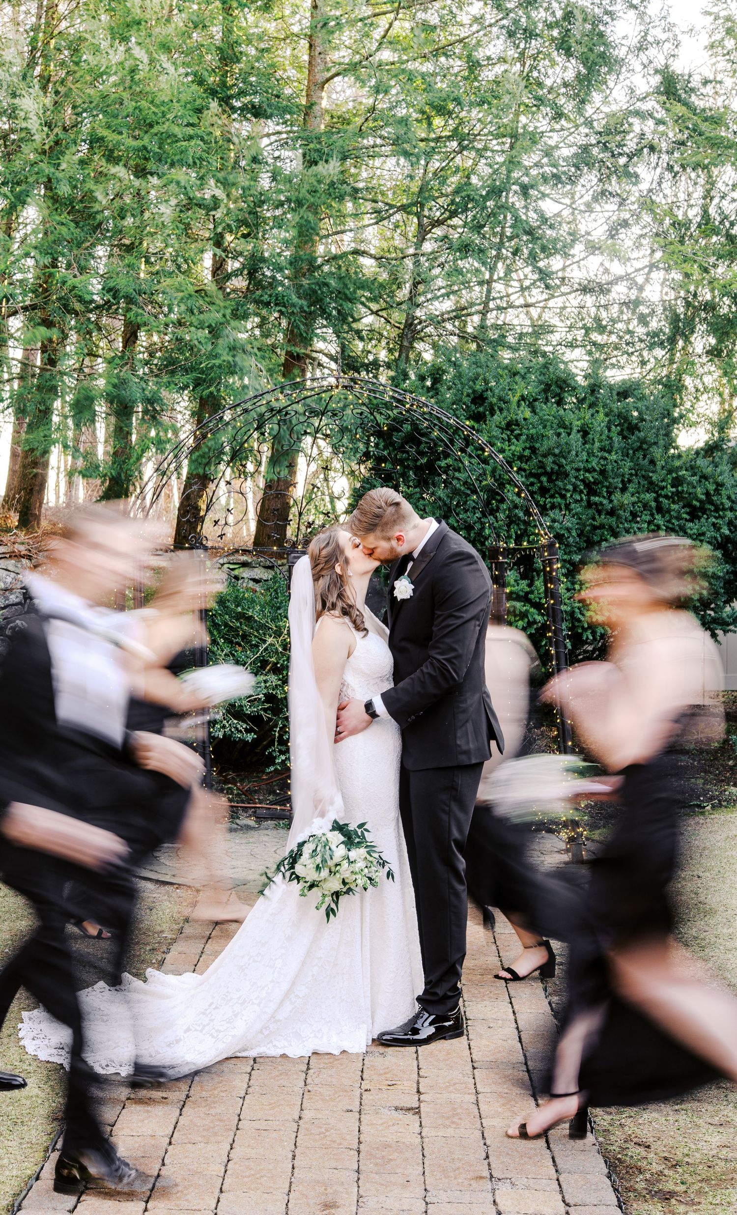 A wedding couple shares an intimate moment on a brick path while blurred figures rush past in the forest background.