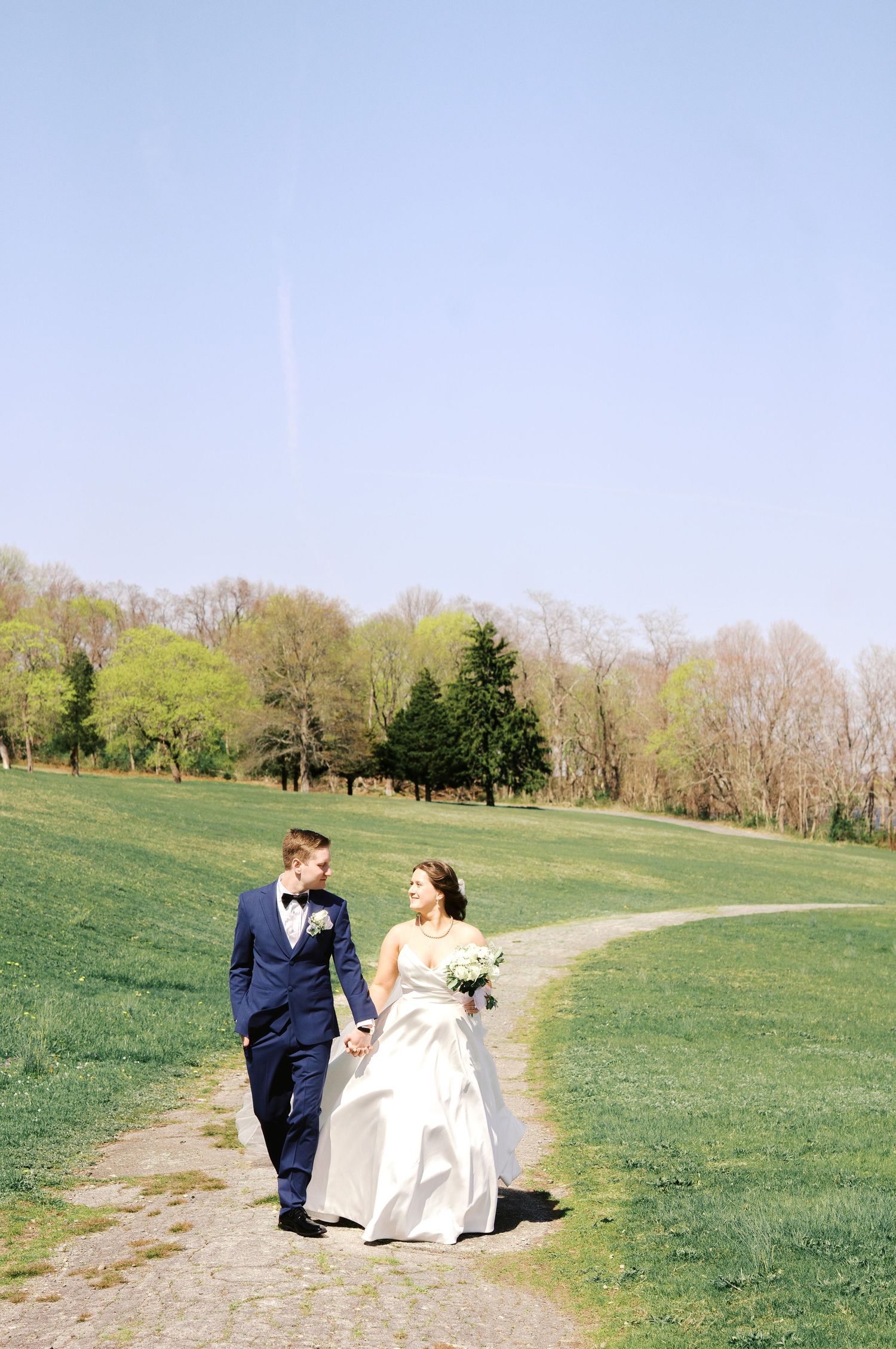 A couple walking along a winding dirt path through a green hillside on their wedding day in springtime.