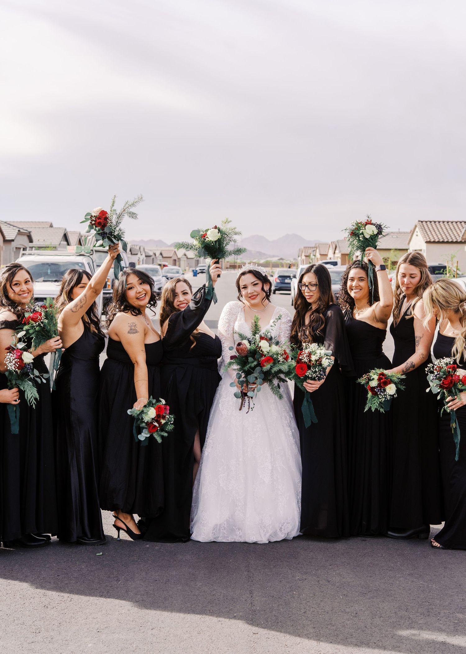 Bridal party wearing elegant black bridesmaid dresses gather together holding red and white floral bouquets for a wedding photo.