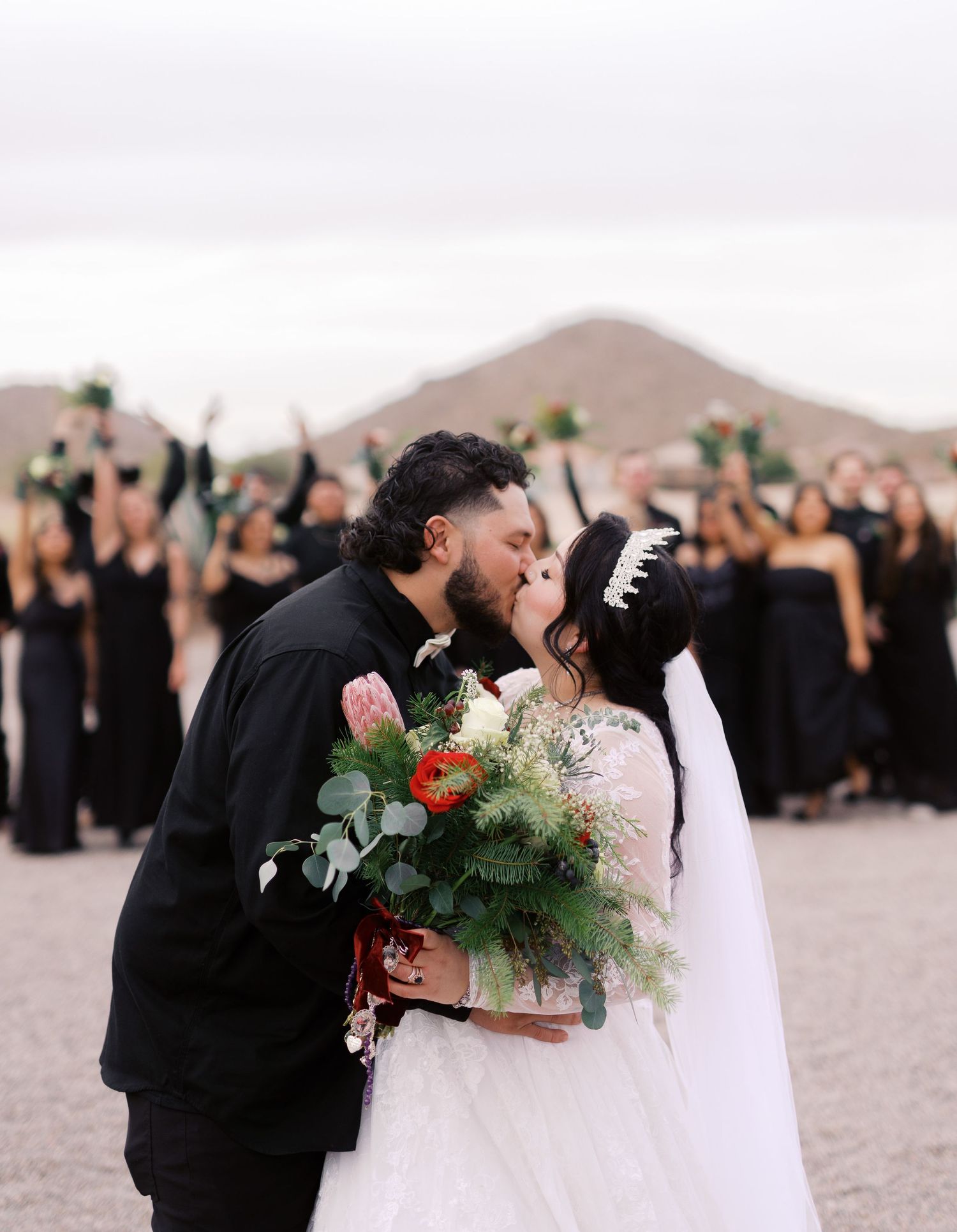 A couple in wedding attire share a kiss while holding a floral bouquet, with their guests in black dresses in the background.