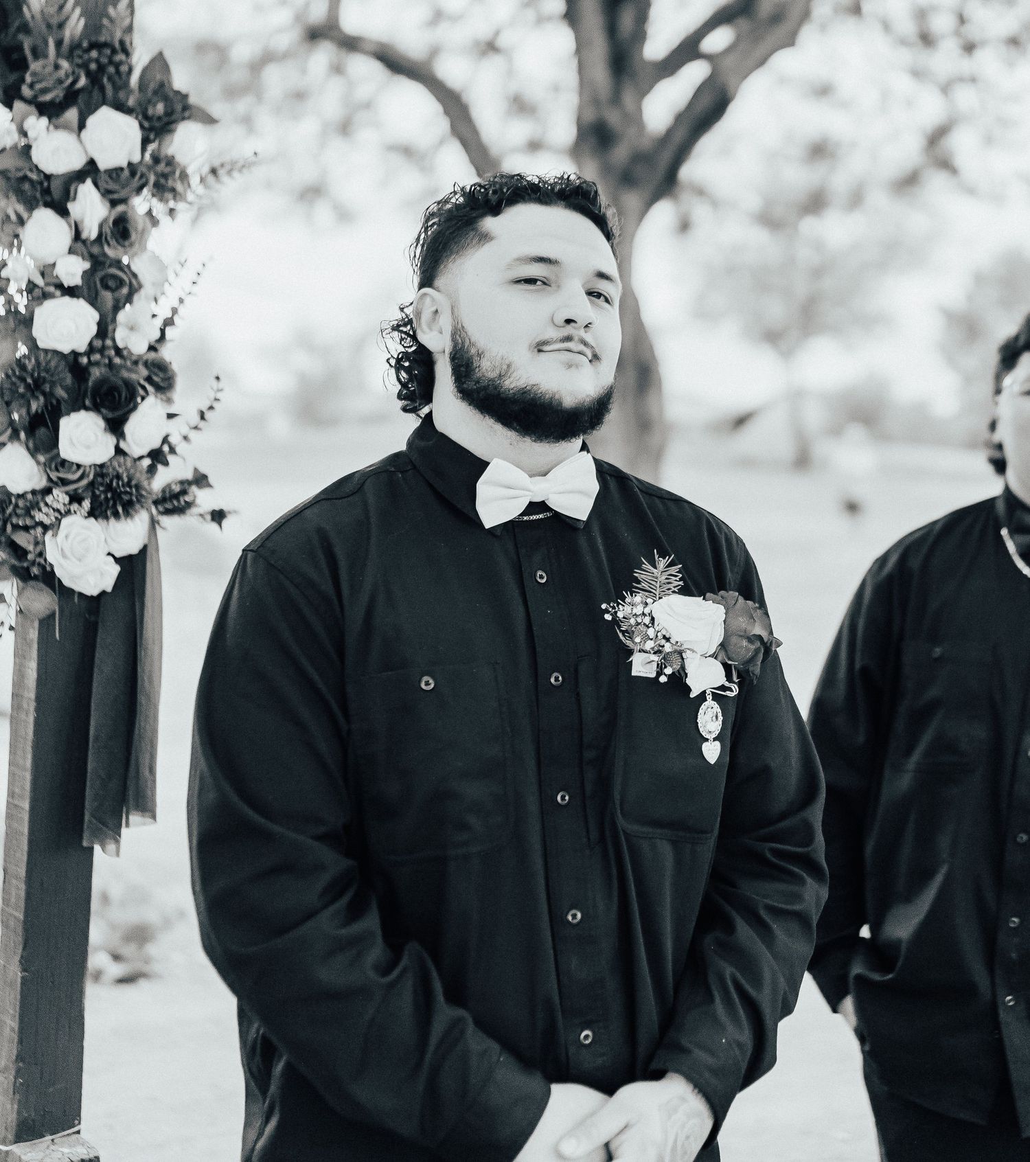Black and white wedding portrait showing a groomsman in a dark button-down shirt with floral boutonniere at an outdoor ceremony.