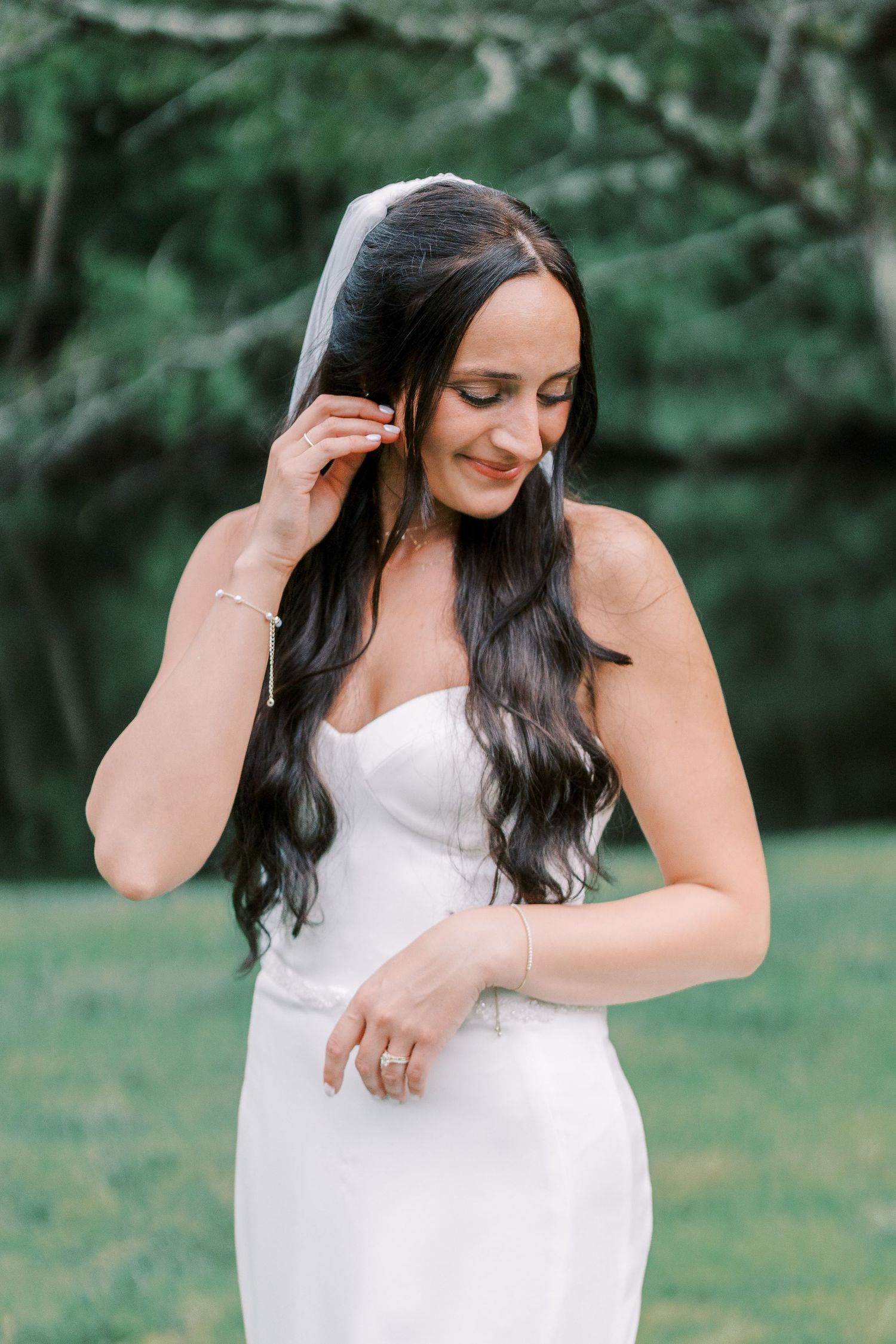 Someone wearing a fitted white strapless wedding dress poses outdoors with long dark hair against a green background.
