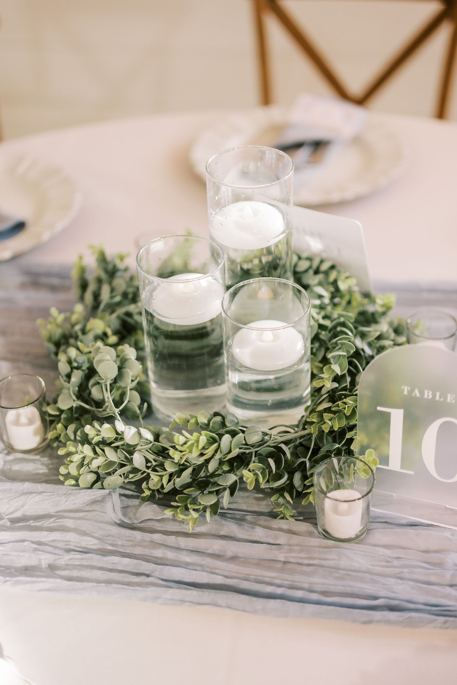 Wedding table centerpiece featuring eucalyptus garland with frosted glass candle holders and white tealights on gray surface.
