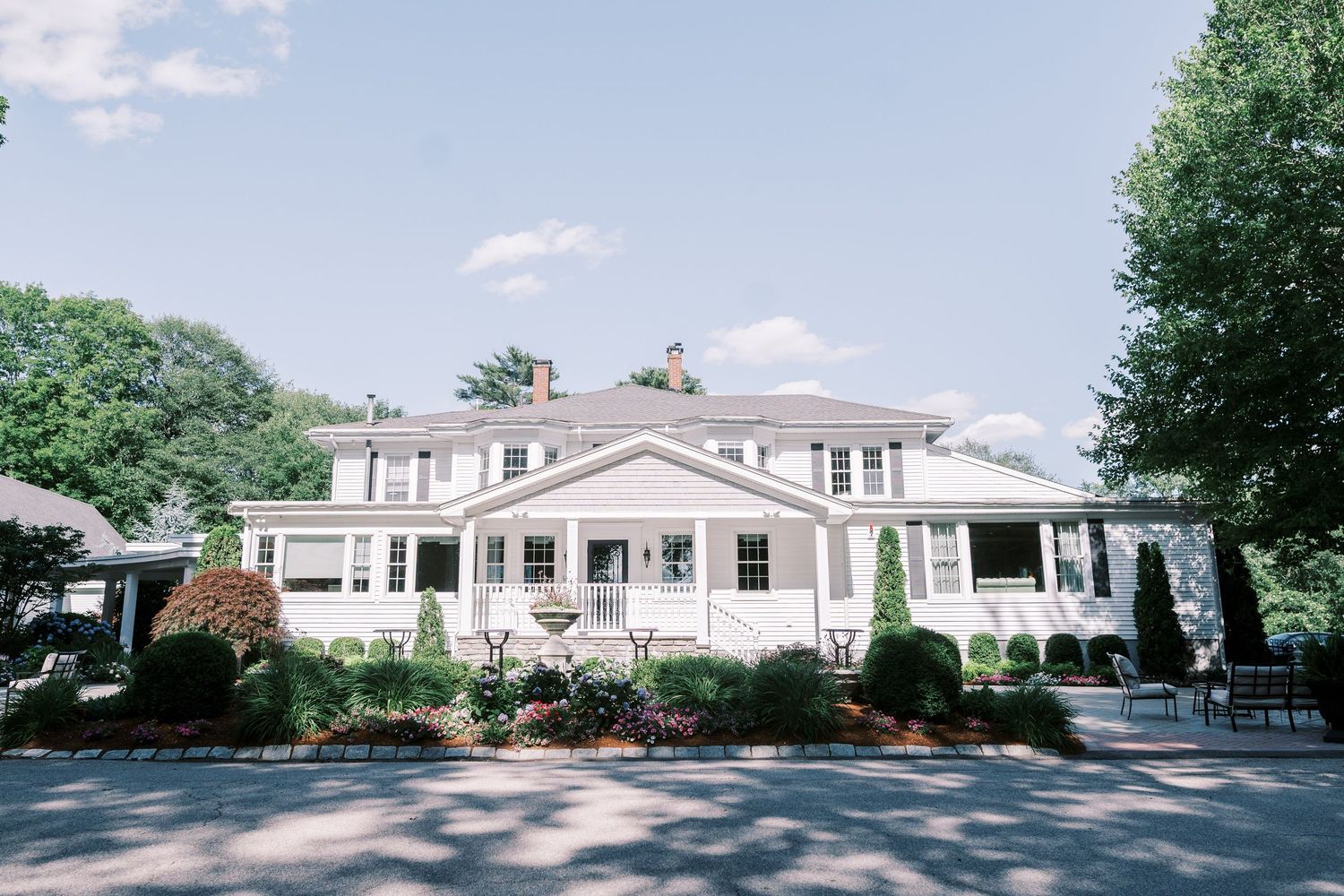Large white colonial style mansion with wraparound porch and manicured landscaping sits along a shaded driveway on a summer day.