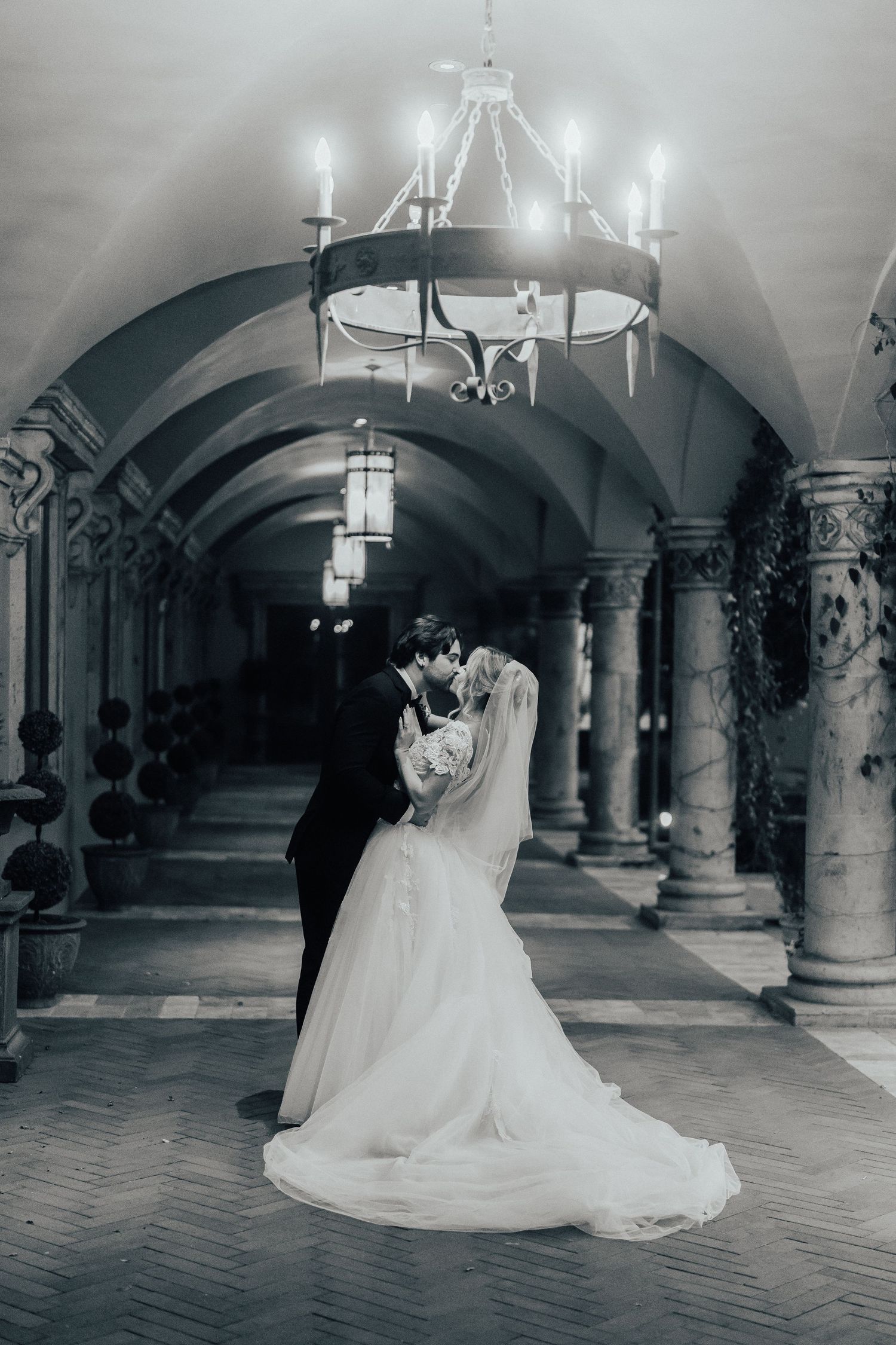 A bride in a flowing white gown twirls gracefully through an arched stone hallway with elegant chandeliers overhead.