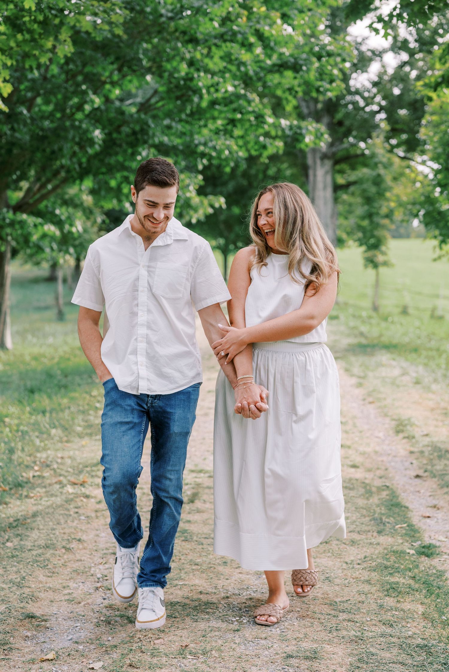 A couple in matching white outfits walking and laughing together on a tree-lined path.