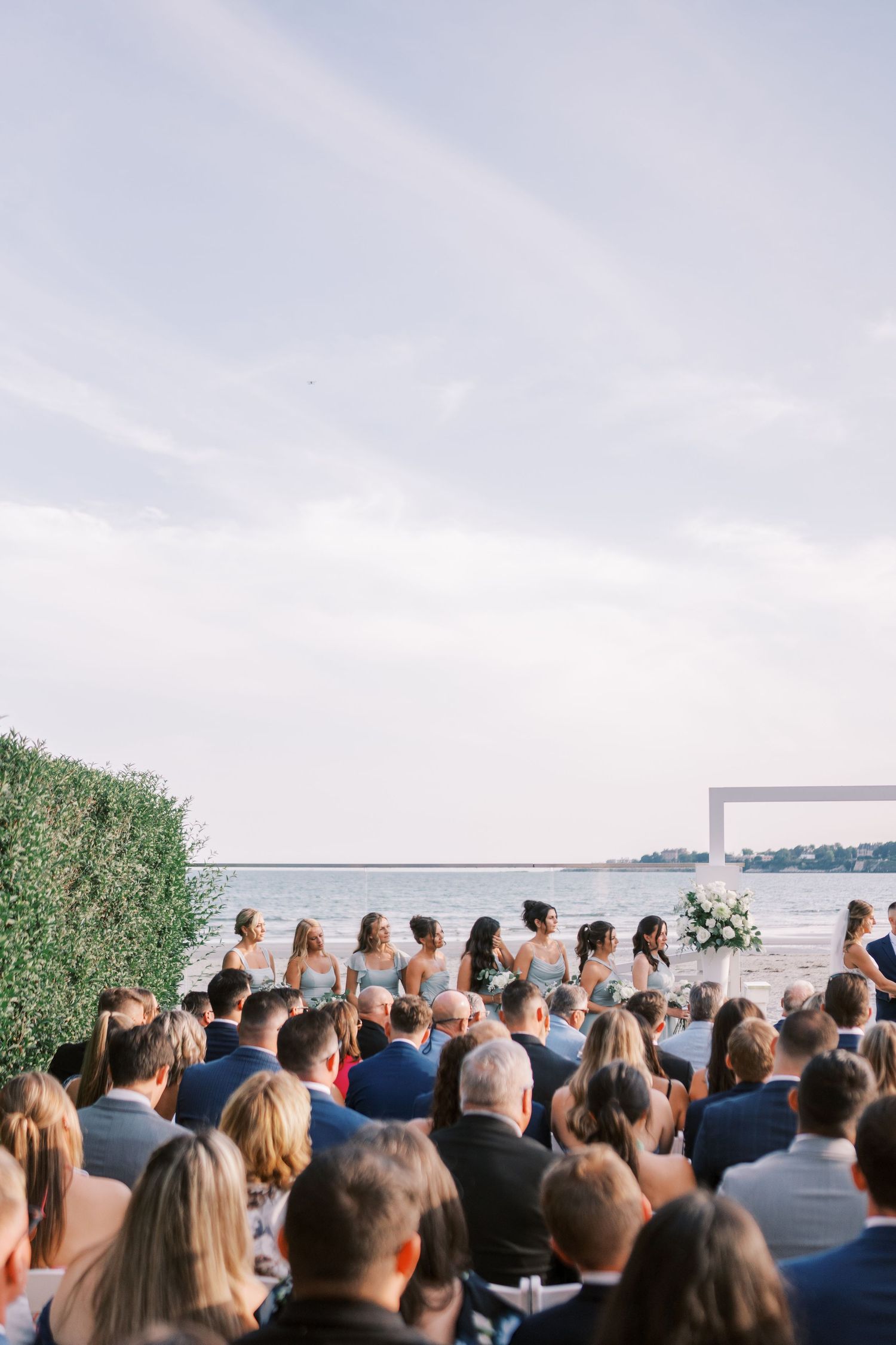 Wedding guests gather for an outdoor waterfront ceremony under cloudy skies with water and greenery in the background.