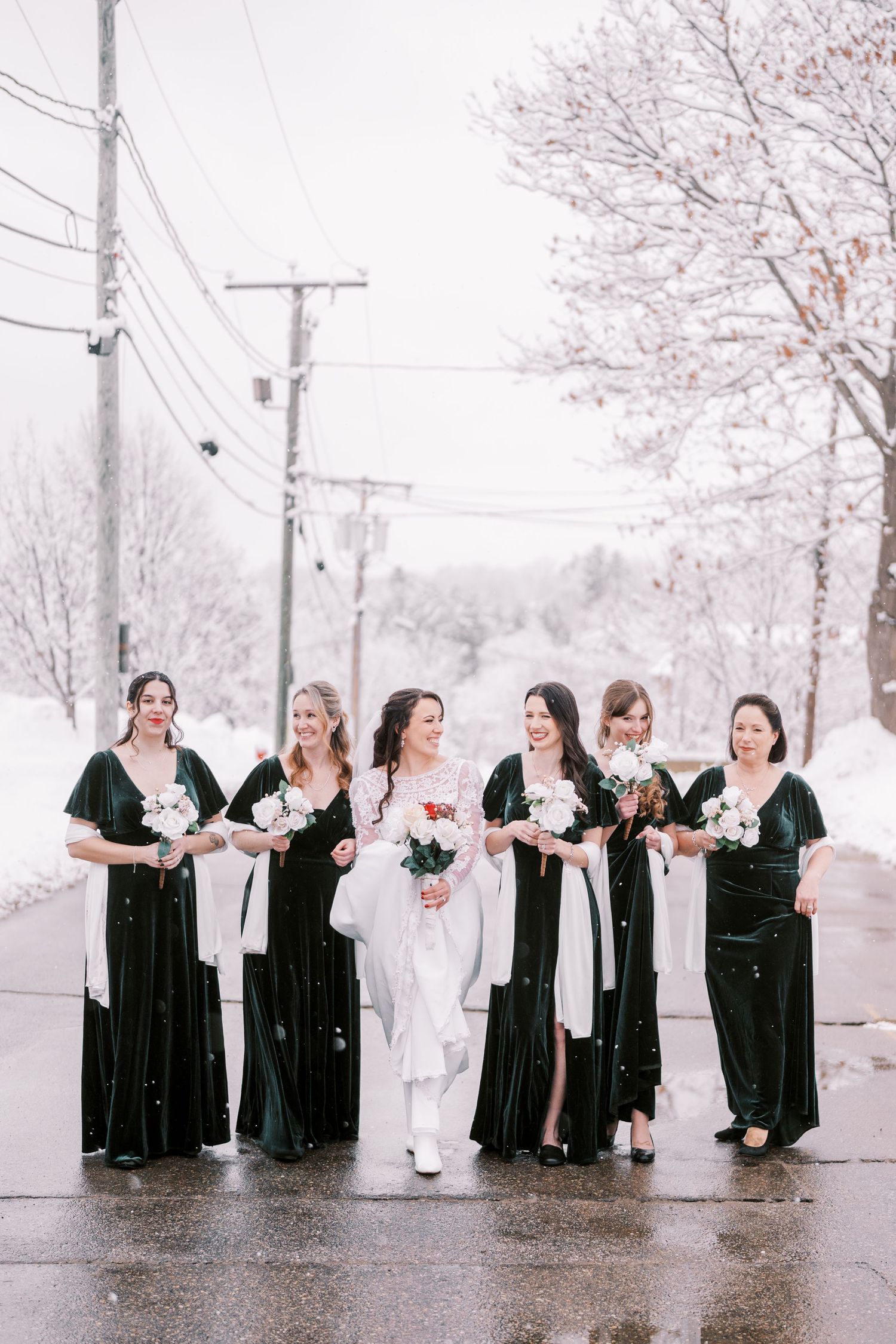 Bridesmaids in elegant black dresses pose with bride in white on snowy winter street.