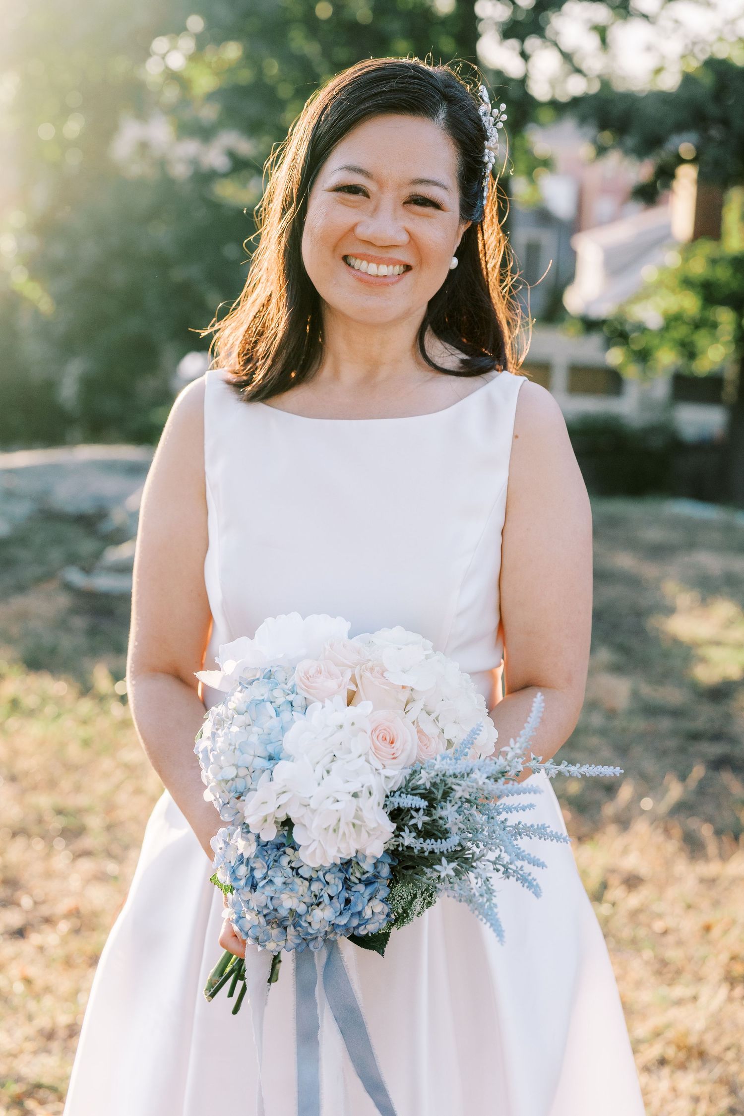 Bride in sleeveless white dress holding delicate blue and white floral bouquet in sunlit setting.