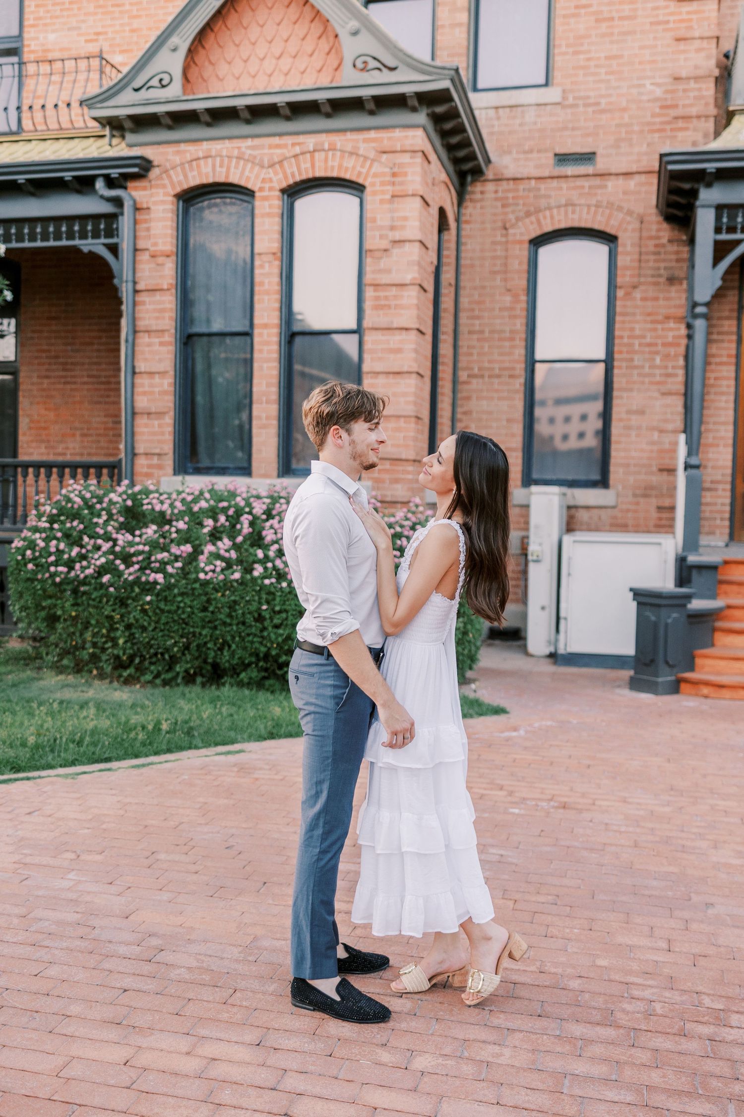 Romantic couple dancing together on brick patio in front of historic building with white dress and casual attire.