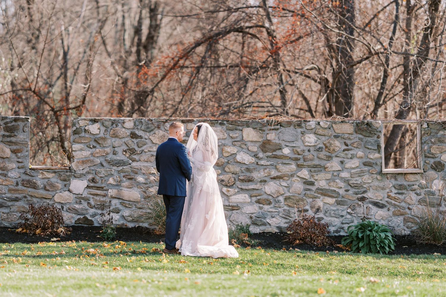 A couple walks along a stone wall in a rustic outdoor setting during an autumn wedding photoshoot.