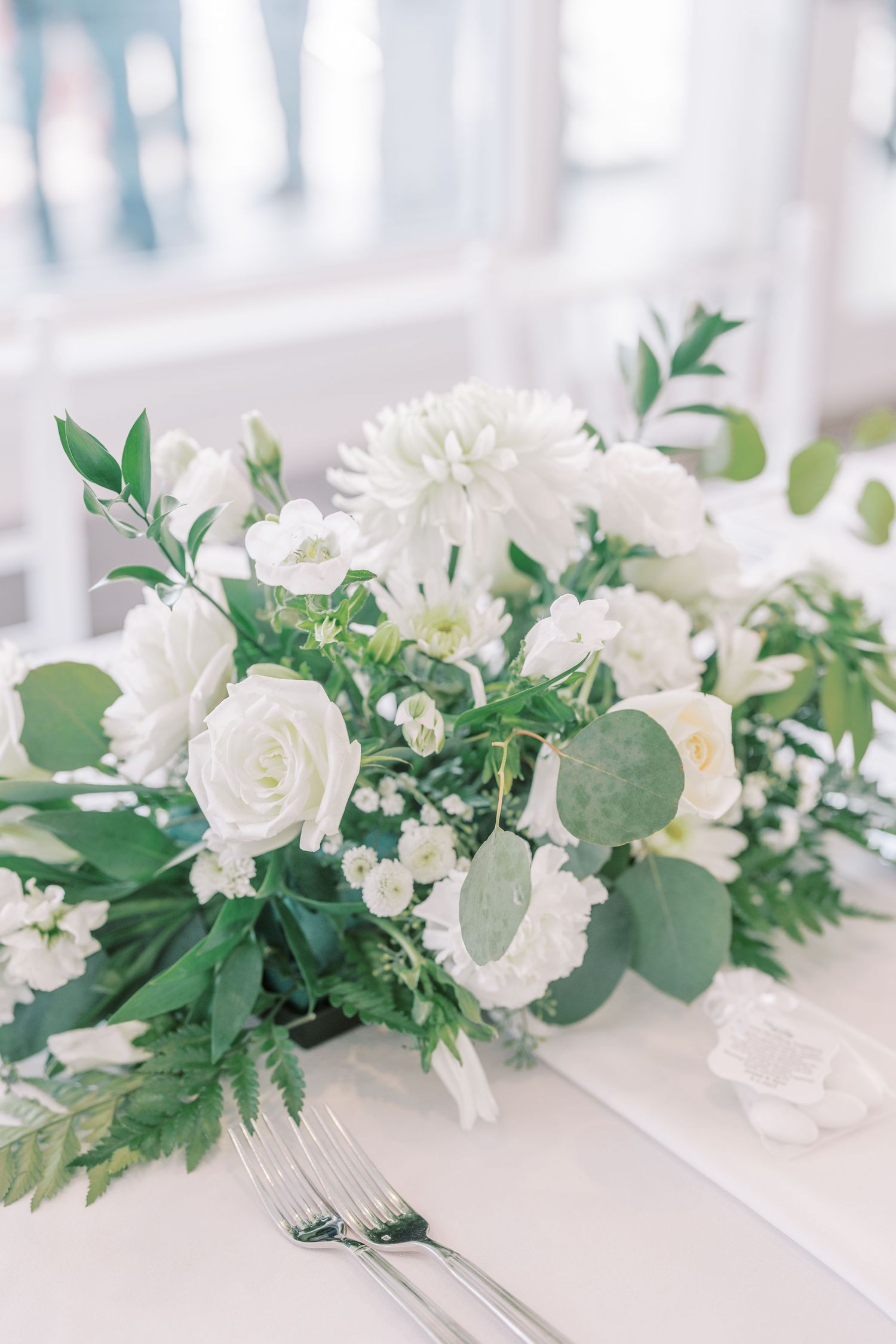 Elegant white rose and eucalyptus wedding centerpiece arrangement on a light table with silverware.