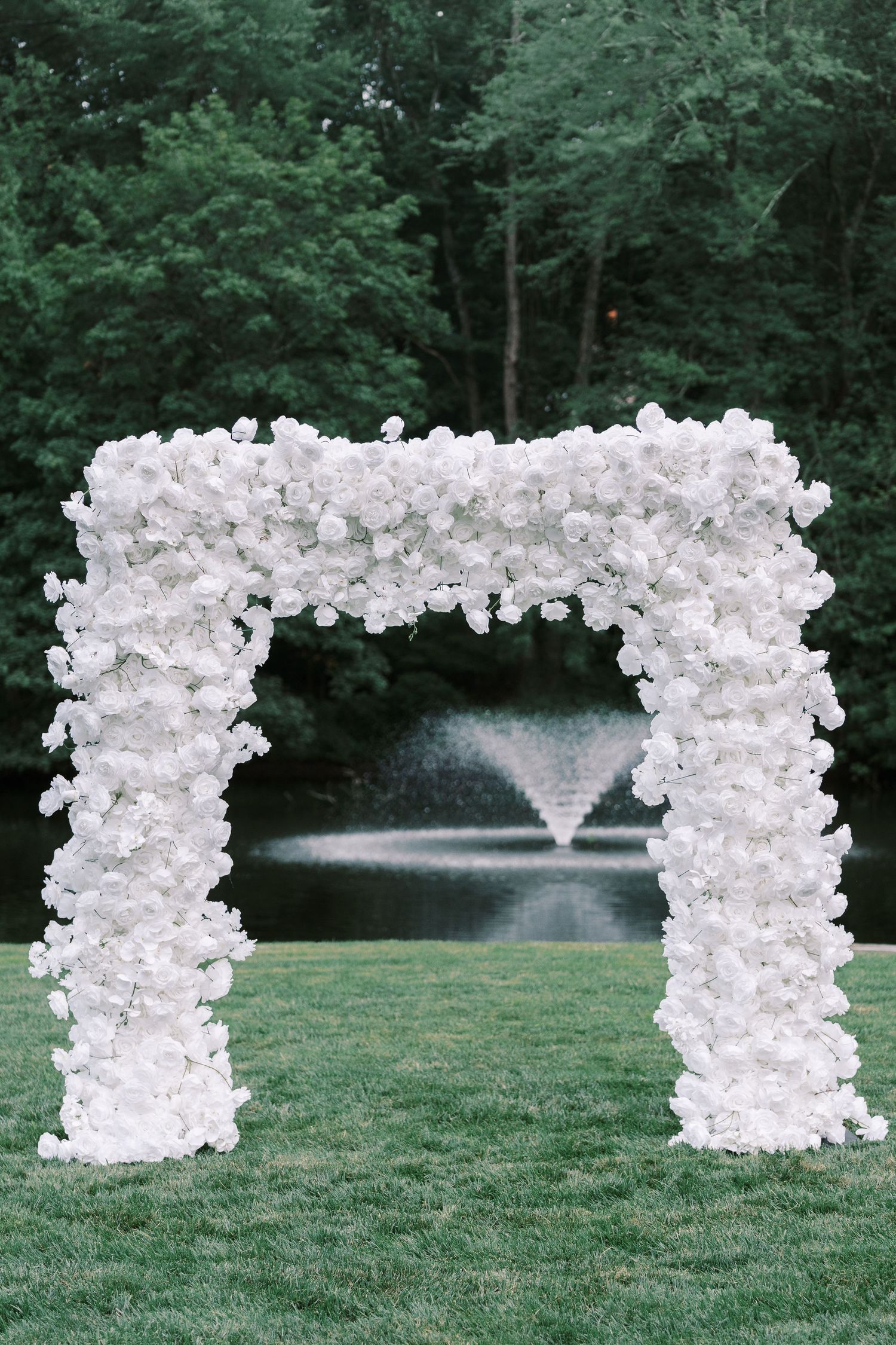 White floral wedding arch with fountain backdrop in lush garden setting.