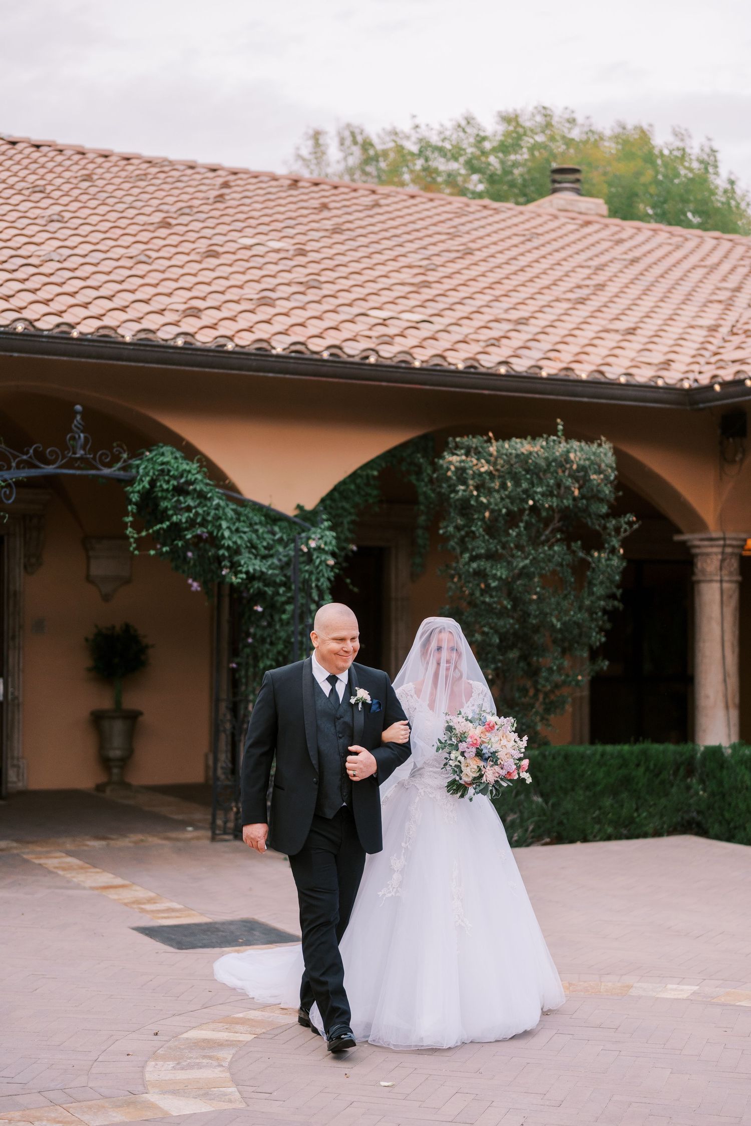 A bride and groom walk together under a Spanish-style covered walkway with terracotta roof tiles and climbing vines.