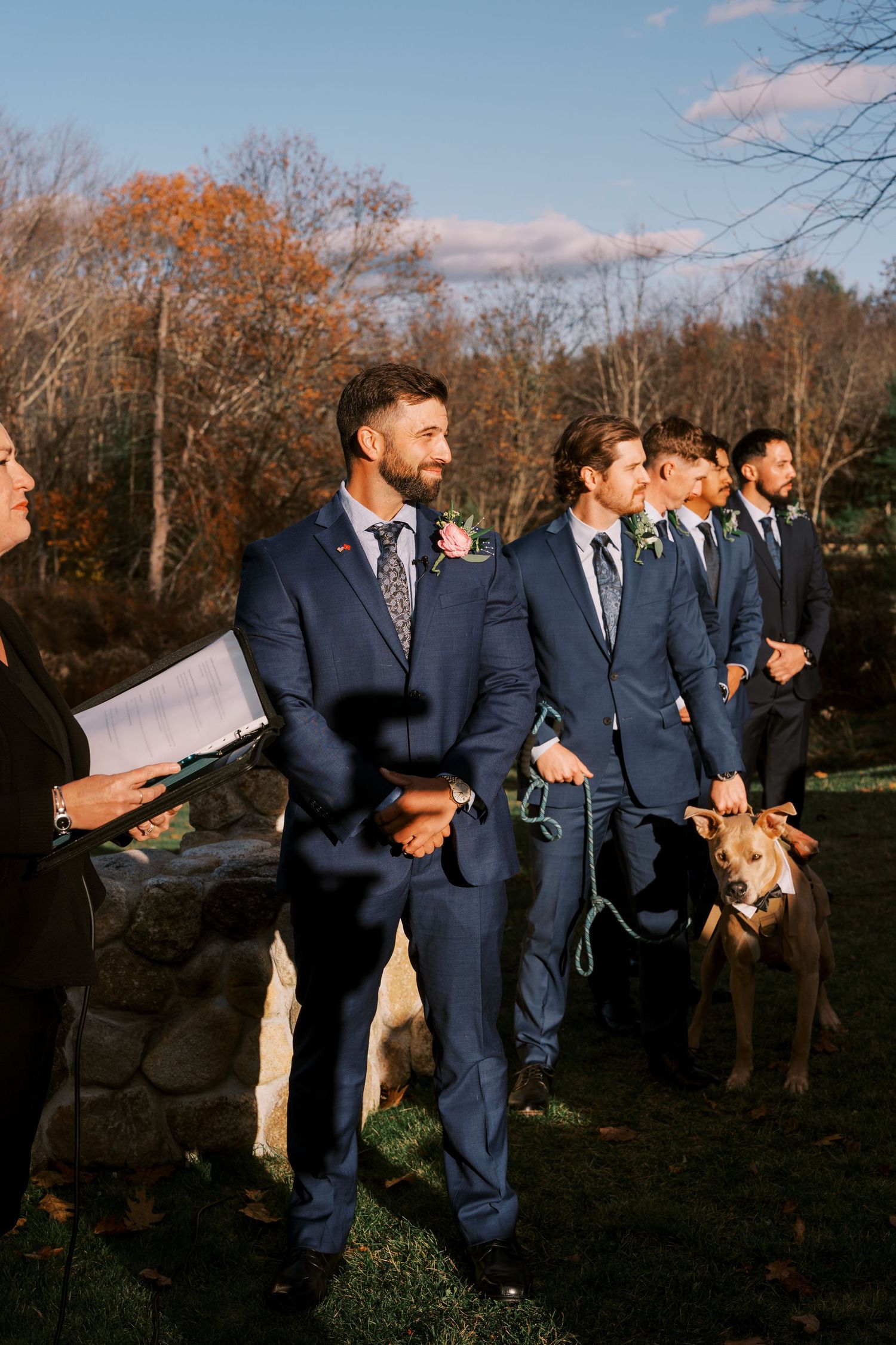 Wedding party in navy blue suits poses outdoors during autumn with bare trees in the background.