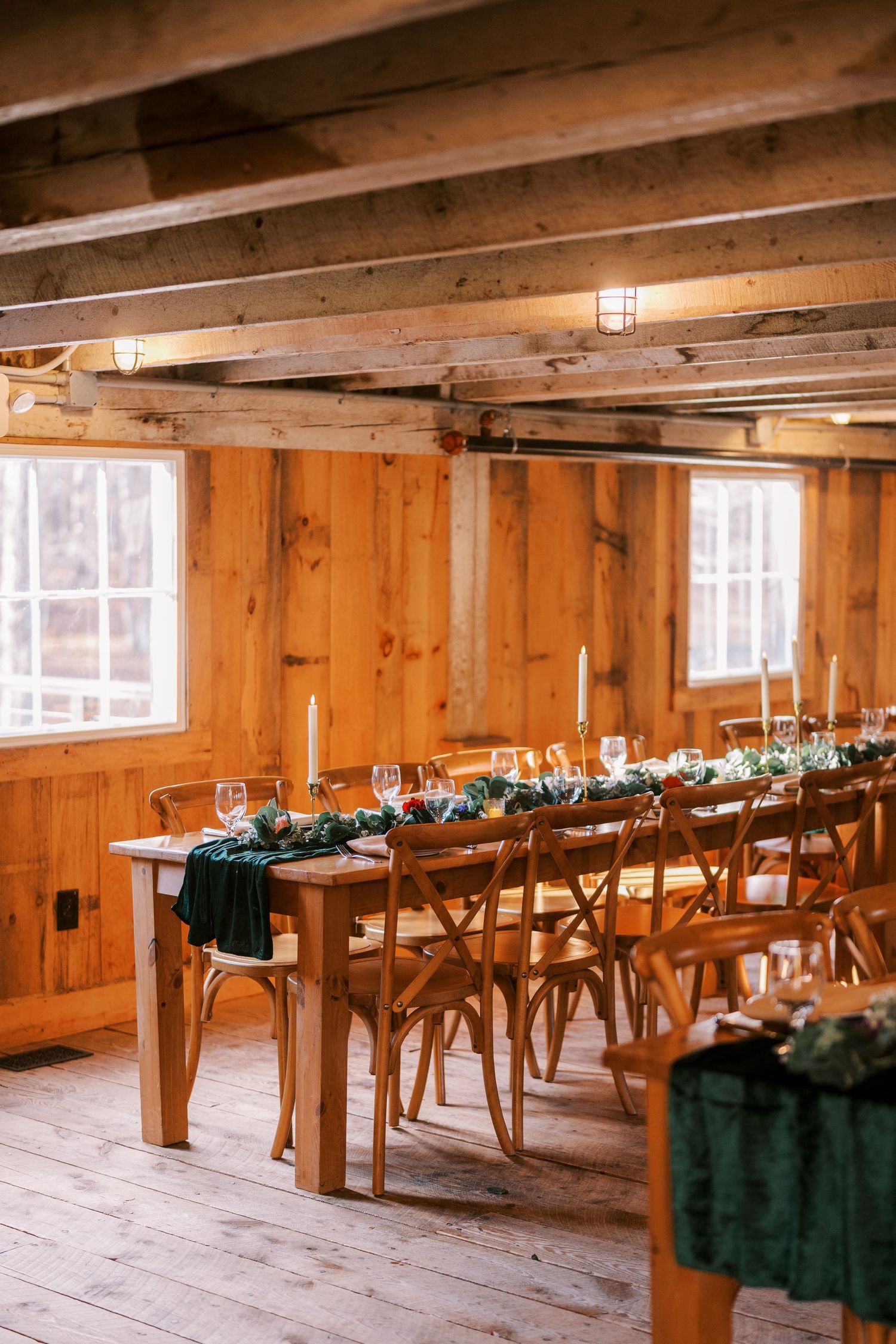 Long wooden farm tables with crossback chairs and dark green linens set up in rustic barn venue with natural lighting.
