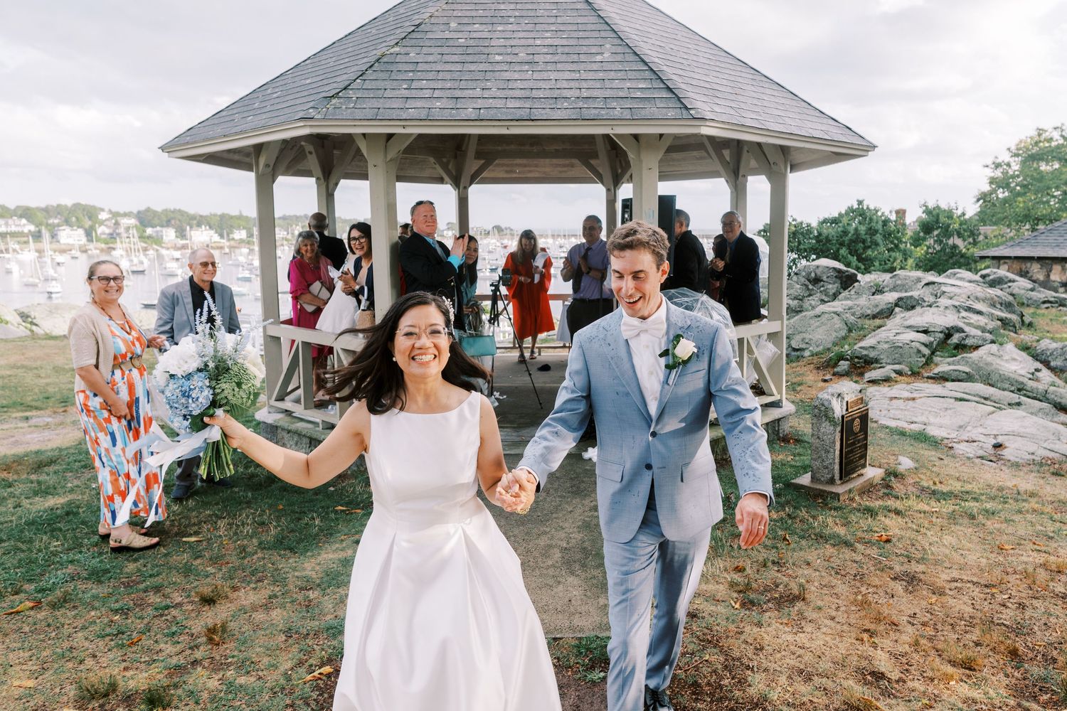 Newlyweds exit a gazebo surrounded by guests at scenic coastal wedding venue.
