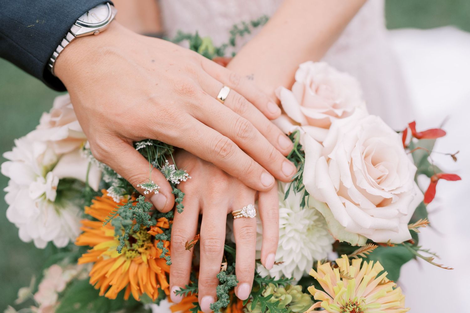 Close-up of hands with wedding rings resting on pink and orange floral bouquet.