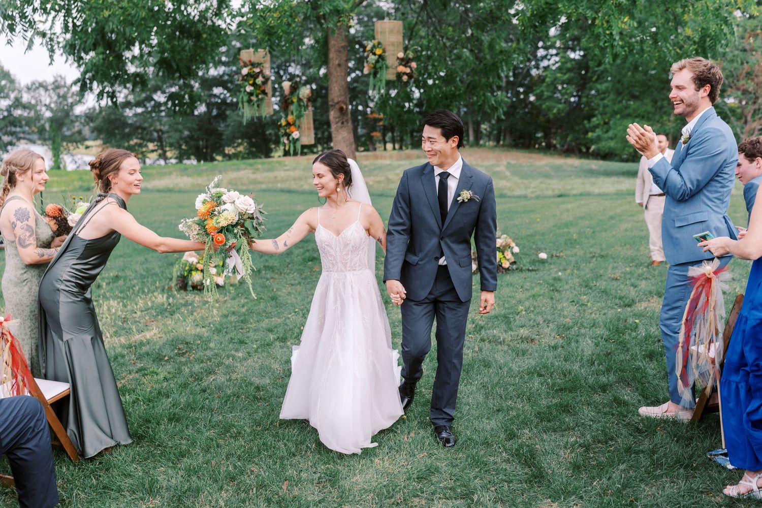 Wedding party members dance and celebrate together on a grassy lawn during an outdoor summer celebration.