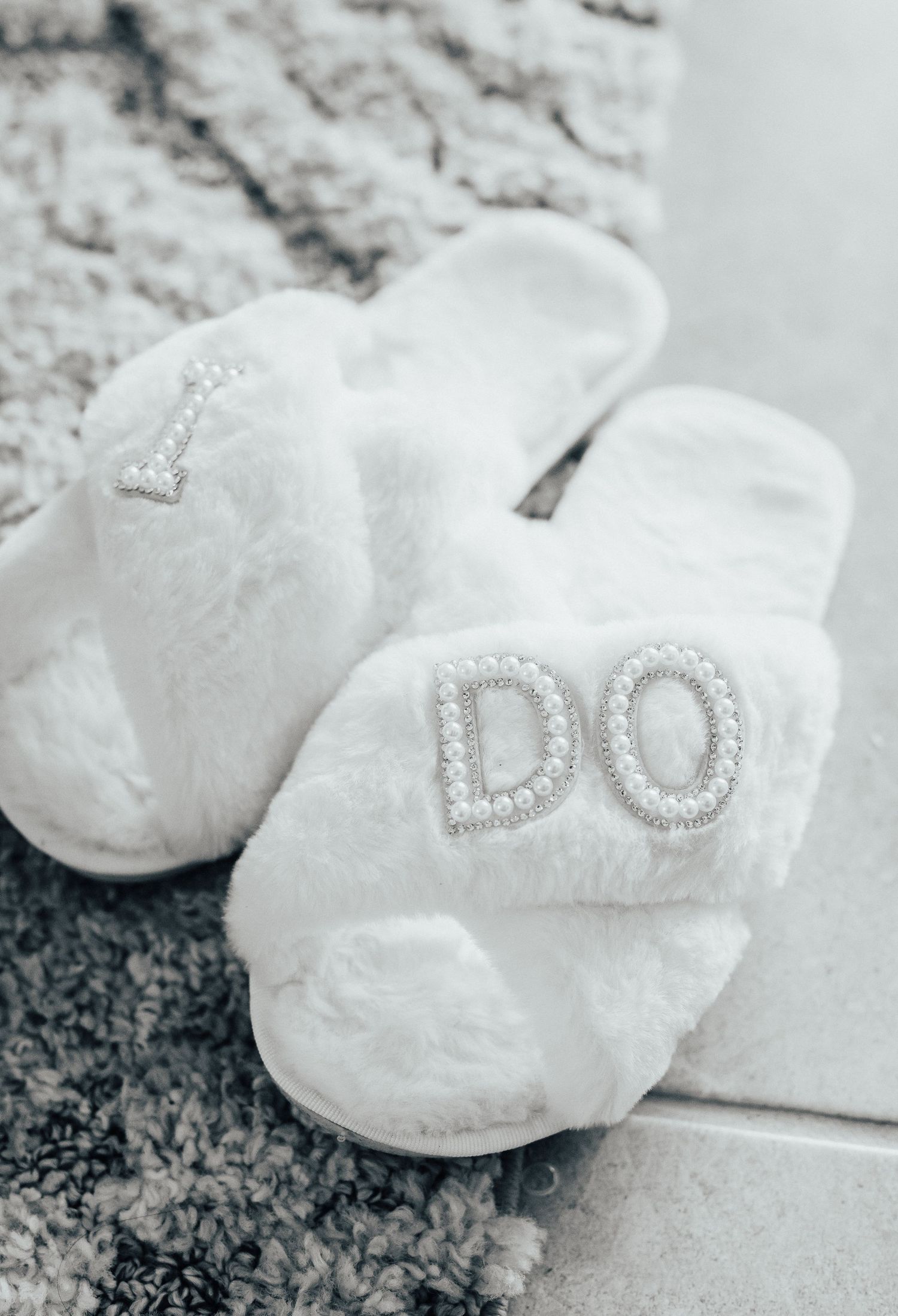 White fuzzy bridal slippers with sparkly 'I DO' text embellishment on a grey textured surface.