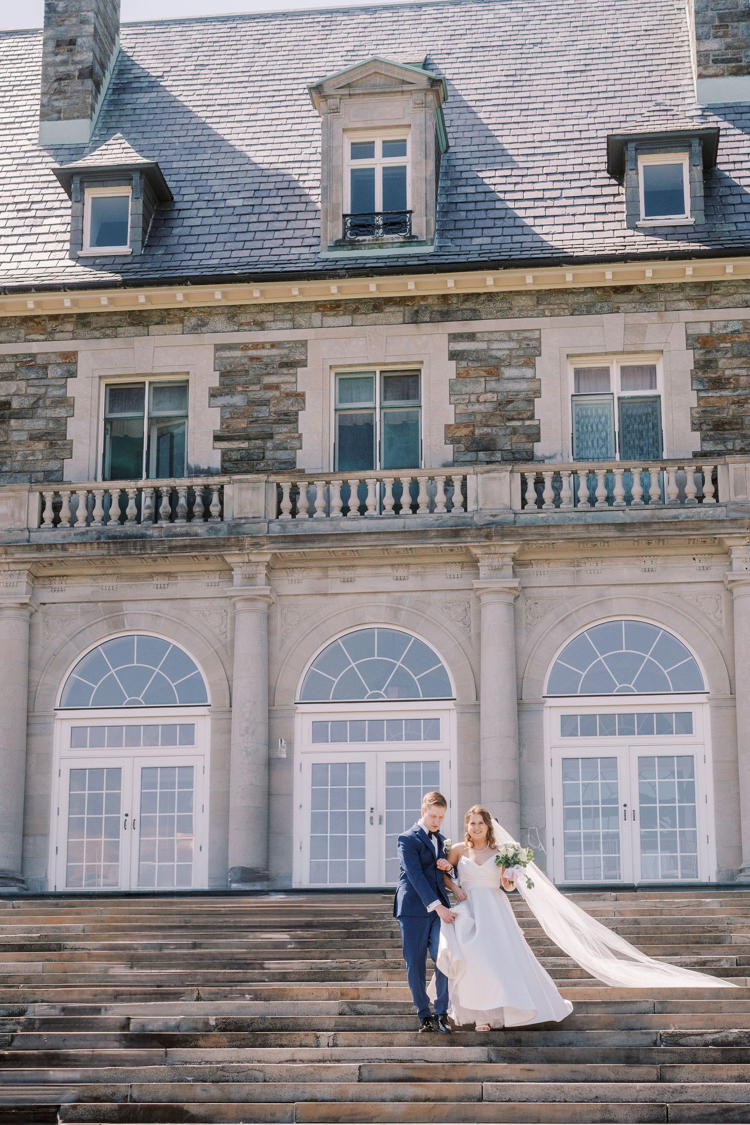 A wedding couple poses on stone steps in front of a grand historic mansion with arched windows.