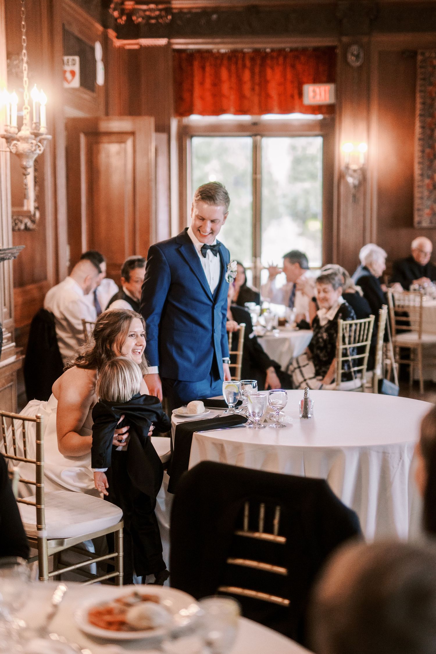 Wedding reception guests enjoying dinner in an elegant wood-paneled ballroom with chandeliers.