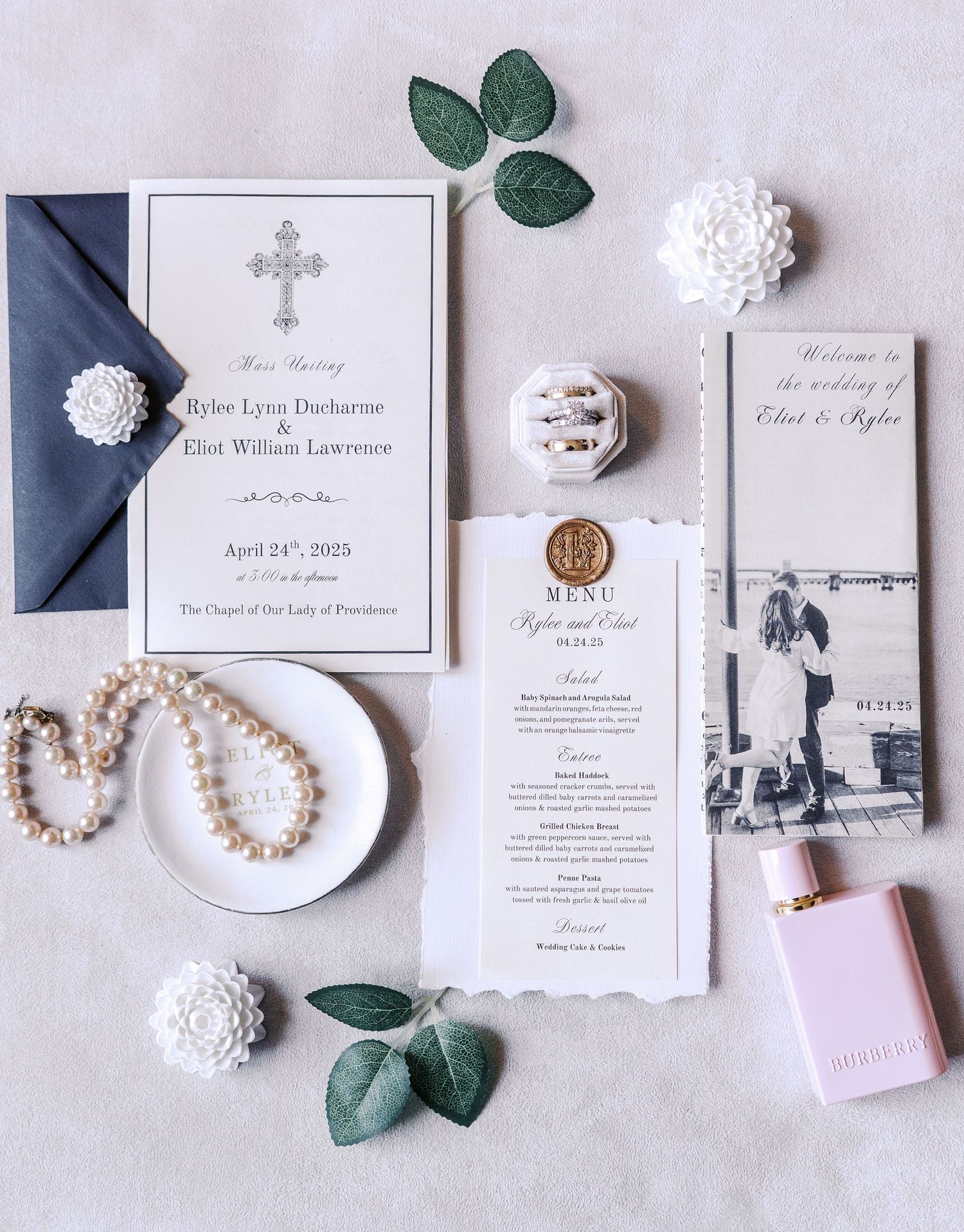 Elegant wedding invitation suite with cross design, navy envelope, pearls, and eucalyptus leaves on light gray background.
