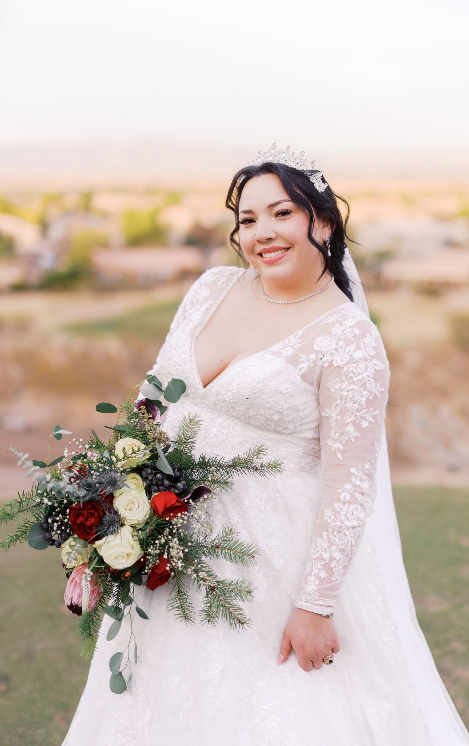 A bride in a white lace wedding dress with long sleeves holds a vibrant winter bouquet of red, white and evergreen florals.