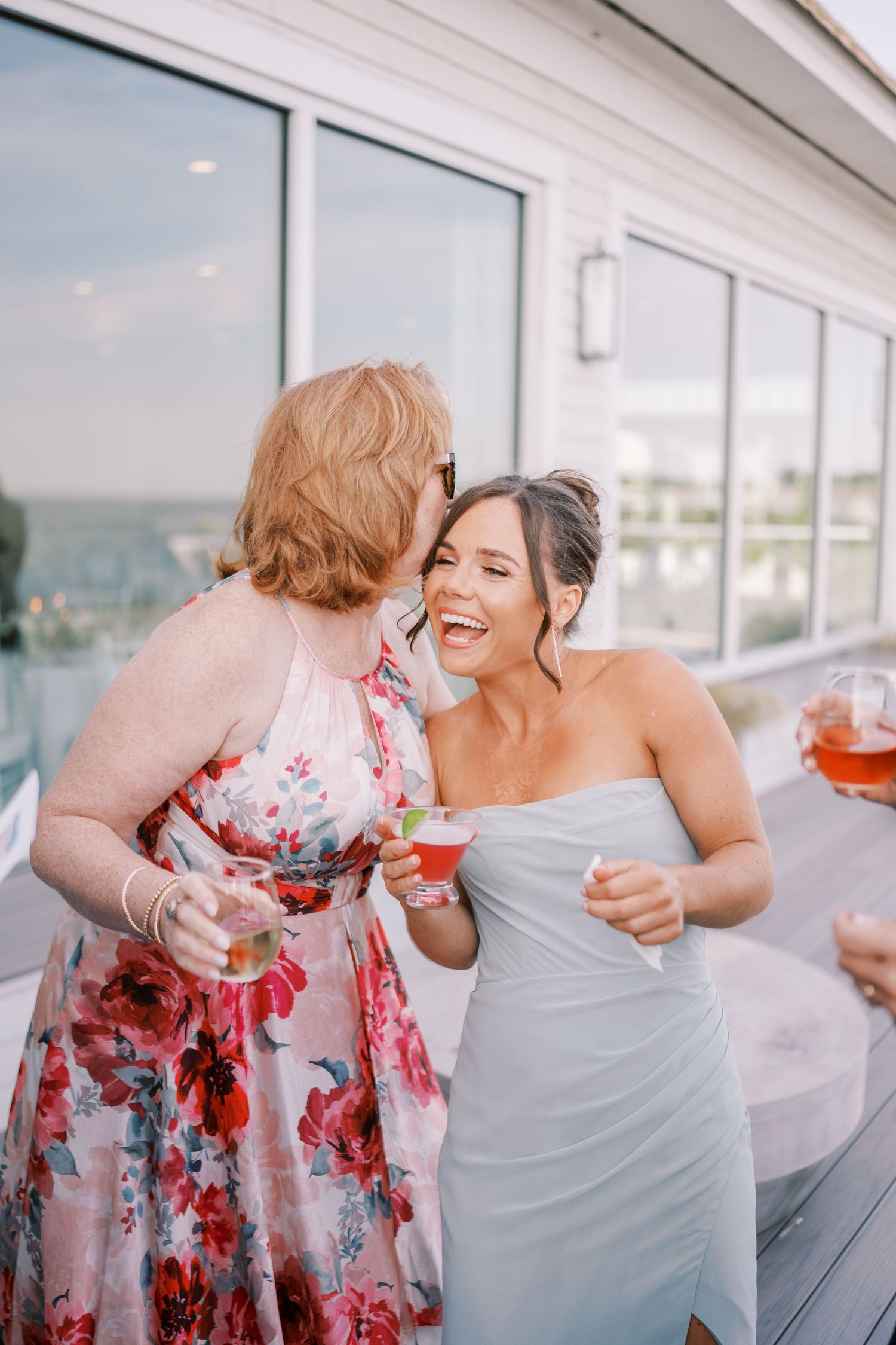 Two people in floral and gray outfits share a joyful moment laughing together on a bright sunlit porch.