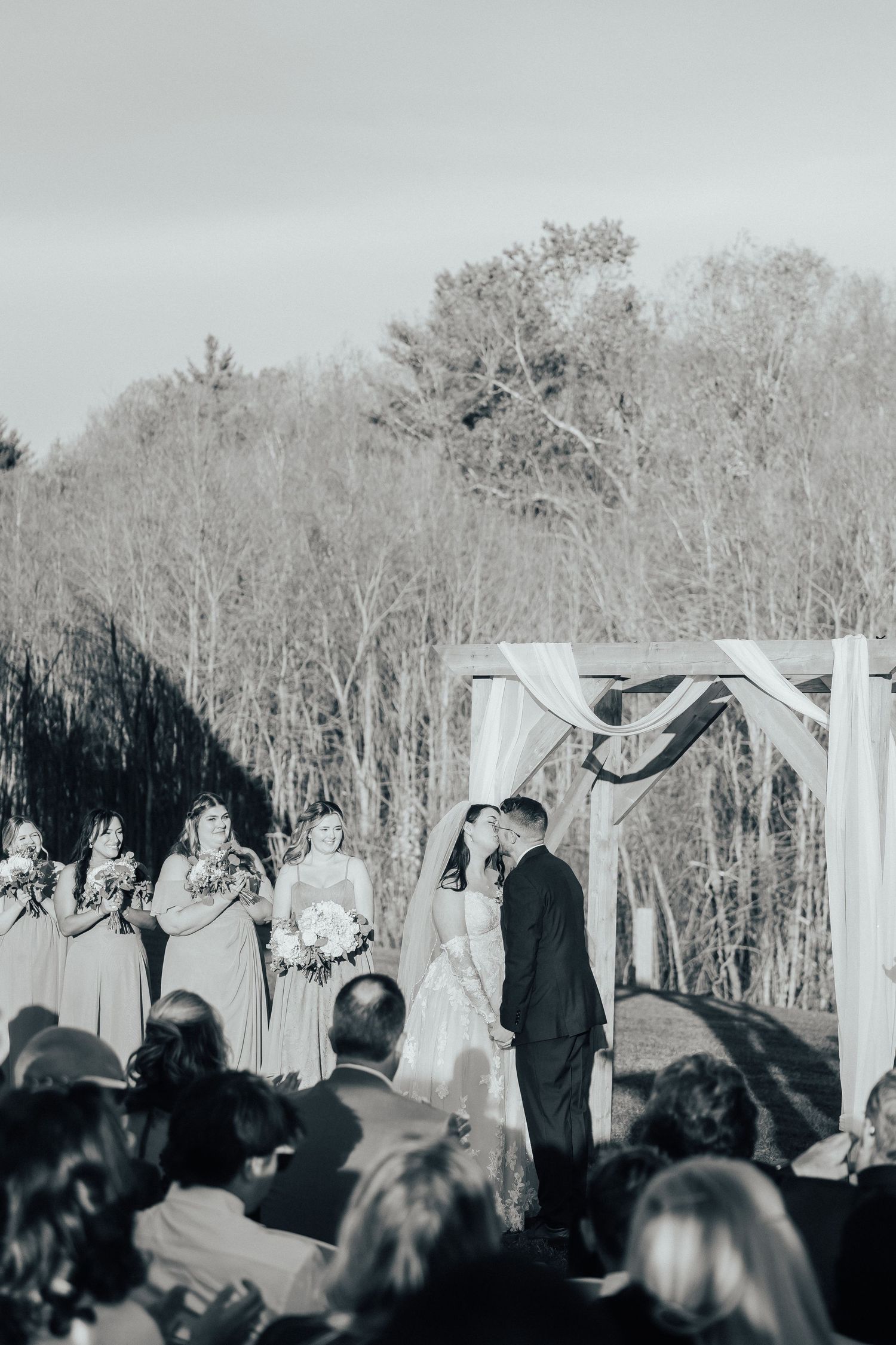 Black and white photo of wedding guests watching an outdoor ceremony with bare winter trees in the background.