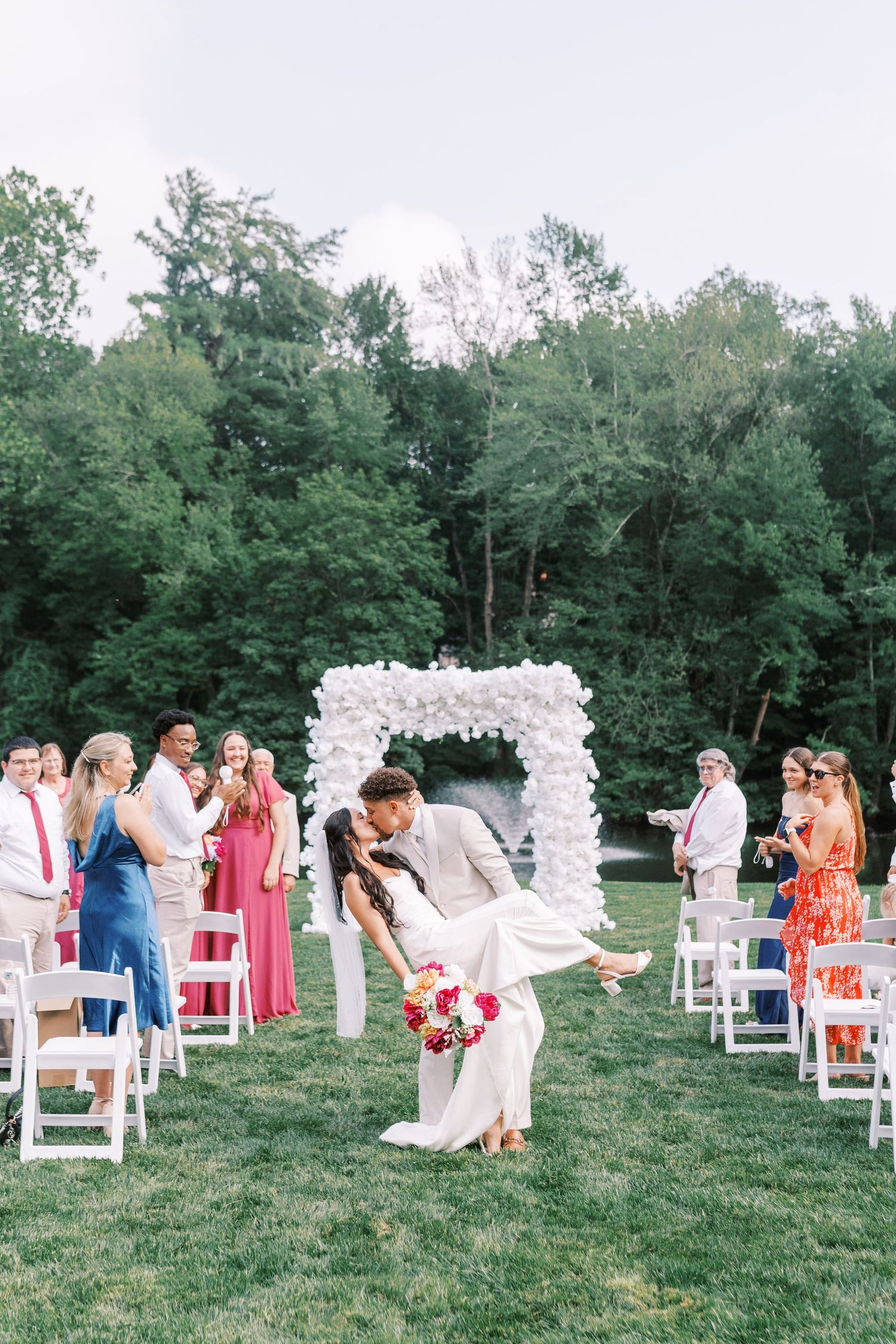 Outdoor wedding ceremony setup with white chairs and floral arch in a garden setting.