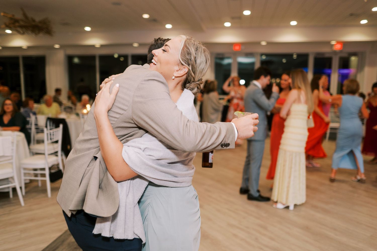 Guests embrace while dancing at an indoor wedding reception with white chairs and other couples dancing in the background.