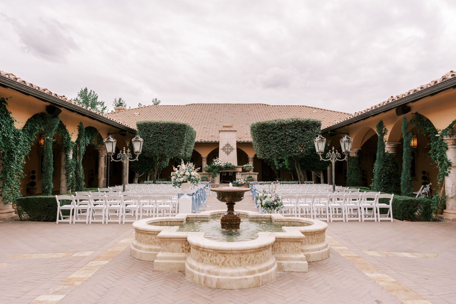 Outdoor wedding ceremony setup with white chairs arranged in rows around a stone fountain in a Mediterranean courtyard.