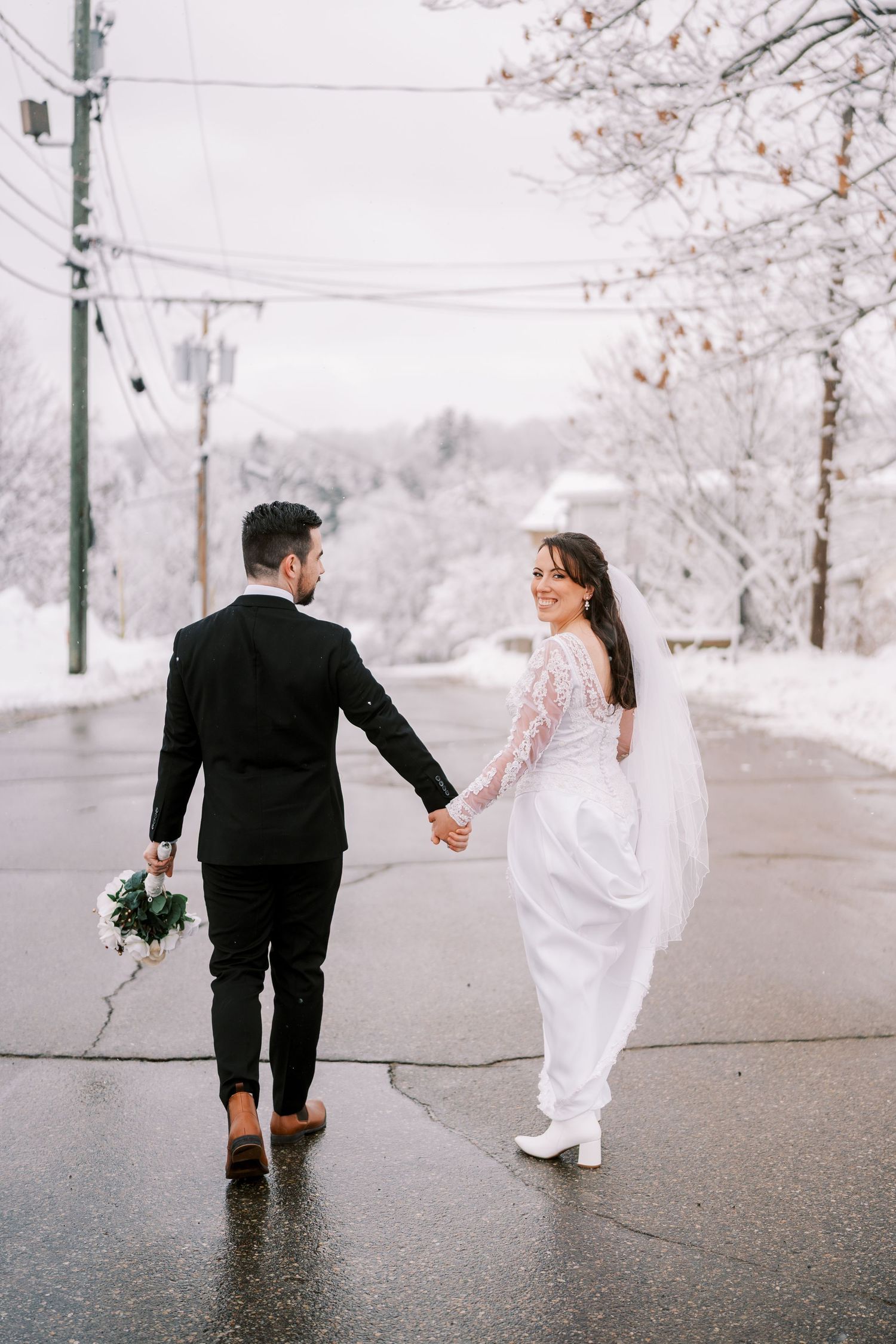 Bride and groom hold hands while walking on wet pavement surrounded by snow-covered trees.