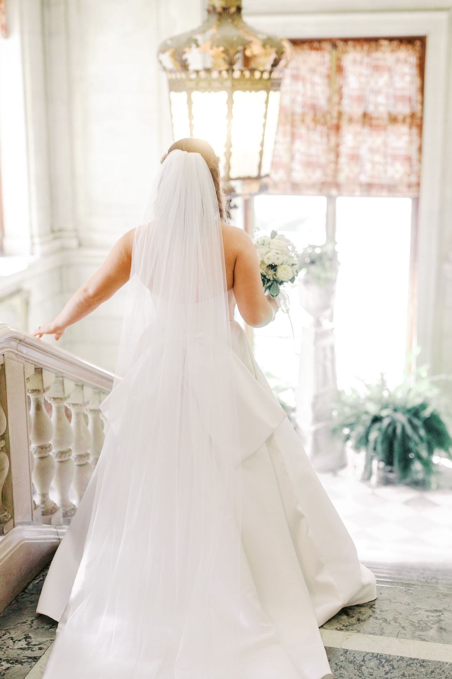 A bride in an elegant white wedding gown and veil stands at the top of a staircase holding a bouquet in soft natural light.