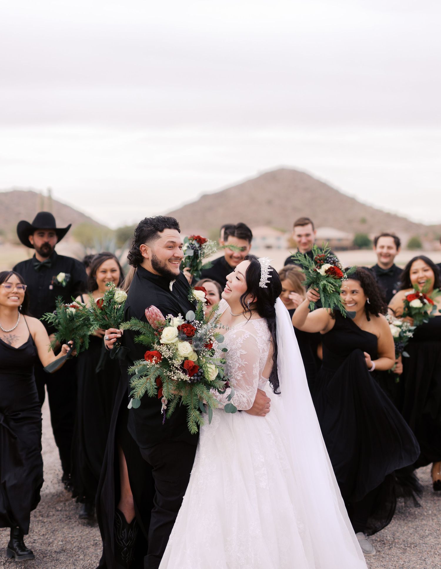 A joyful wedding party in black dresses and formal attire poses together against a desert mountain landscape.