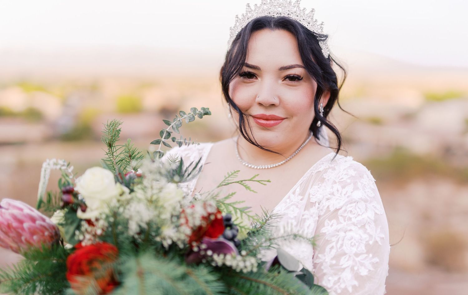 A bride in lace dress smiles while holding an elegant bouquet with white and red flowers against a desert landscape.